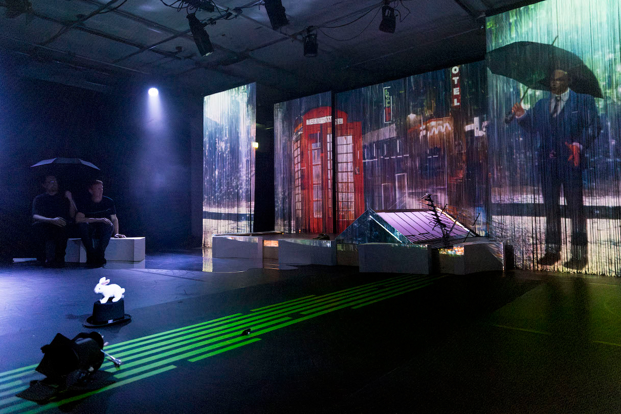 Eine atmosphärische Theaterszene mit einer britischen roten Telefonzelle als Projektion im Hintergrund. Zwei Personen mit Regenschirmen sitzen links im Bild auf weißen Podesten. Ein weißer, beleuchteter Hase ist im Vordergrund platziert. Grüne Laserlinien durchziehen den Bühnenboden. Die Szene wird von einem einzelnen Spotlight beleuchtet und vermittelt eine regnerische London-Atmosphäre.
