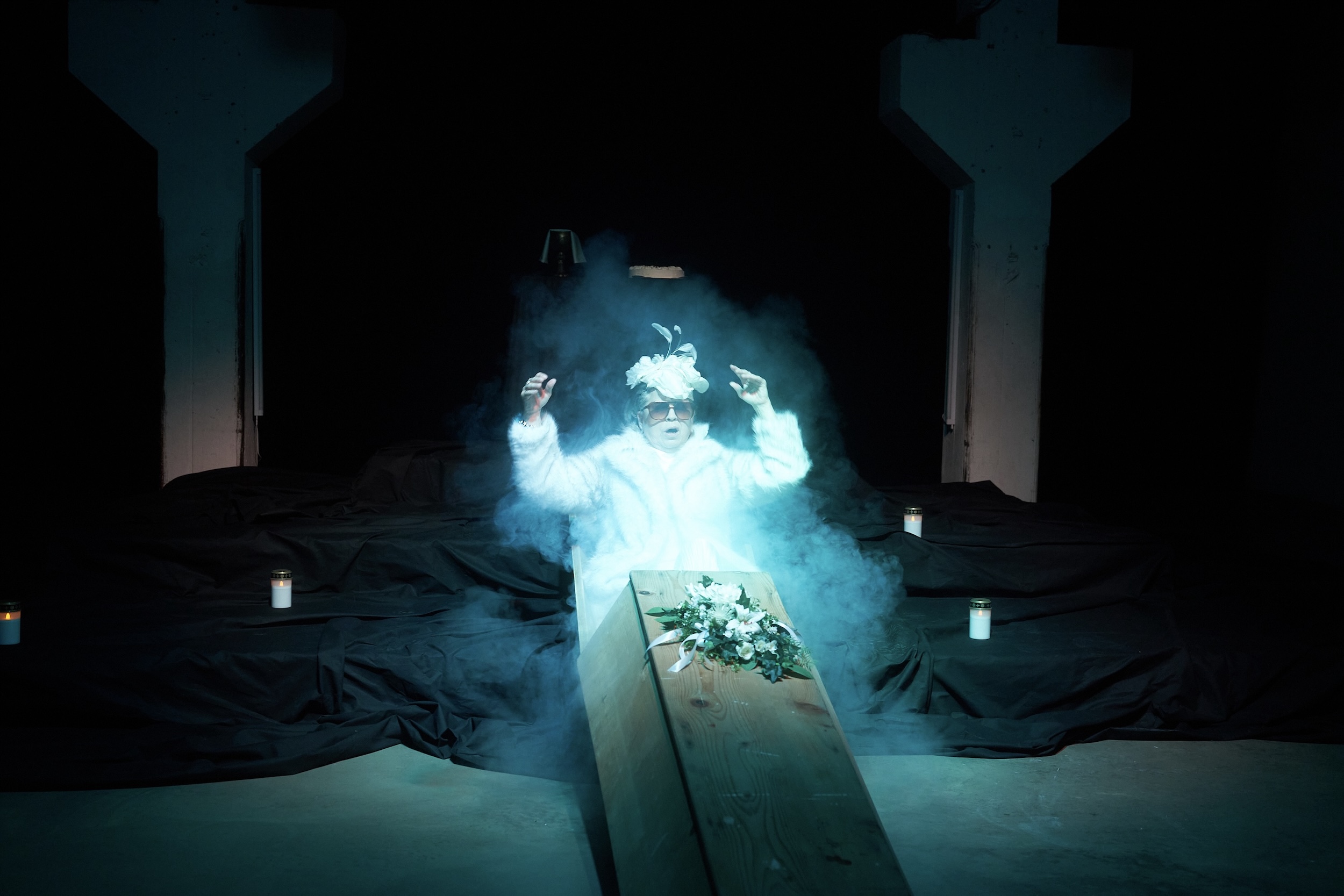 Person in weißem Kunstpelzmantel und Kopfschmuck sitzt aufrecht aus einem offenen Sarg mit Blumenkranz auf dem Deckel auf, umgeben von grünlichem Theaternebel, im dunklen Raum mit vereinzelten Grablichtern.