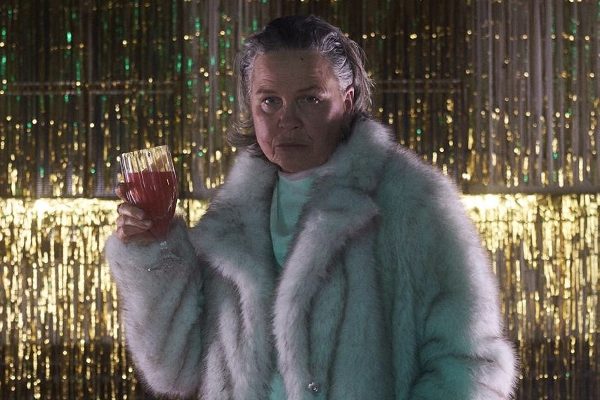 Person in hellem Kunstpelzmantel hält ein Glas mit rotem Getränk in der Hand, steht vor golden glitzerndem Vorhang in bühnenartiger Atmosphäre; das Gesicht ist unkenntlich gemacht.