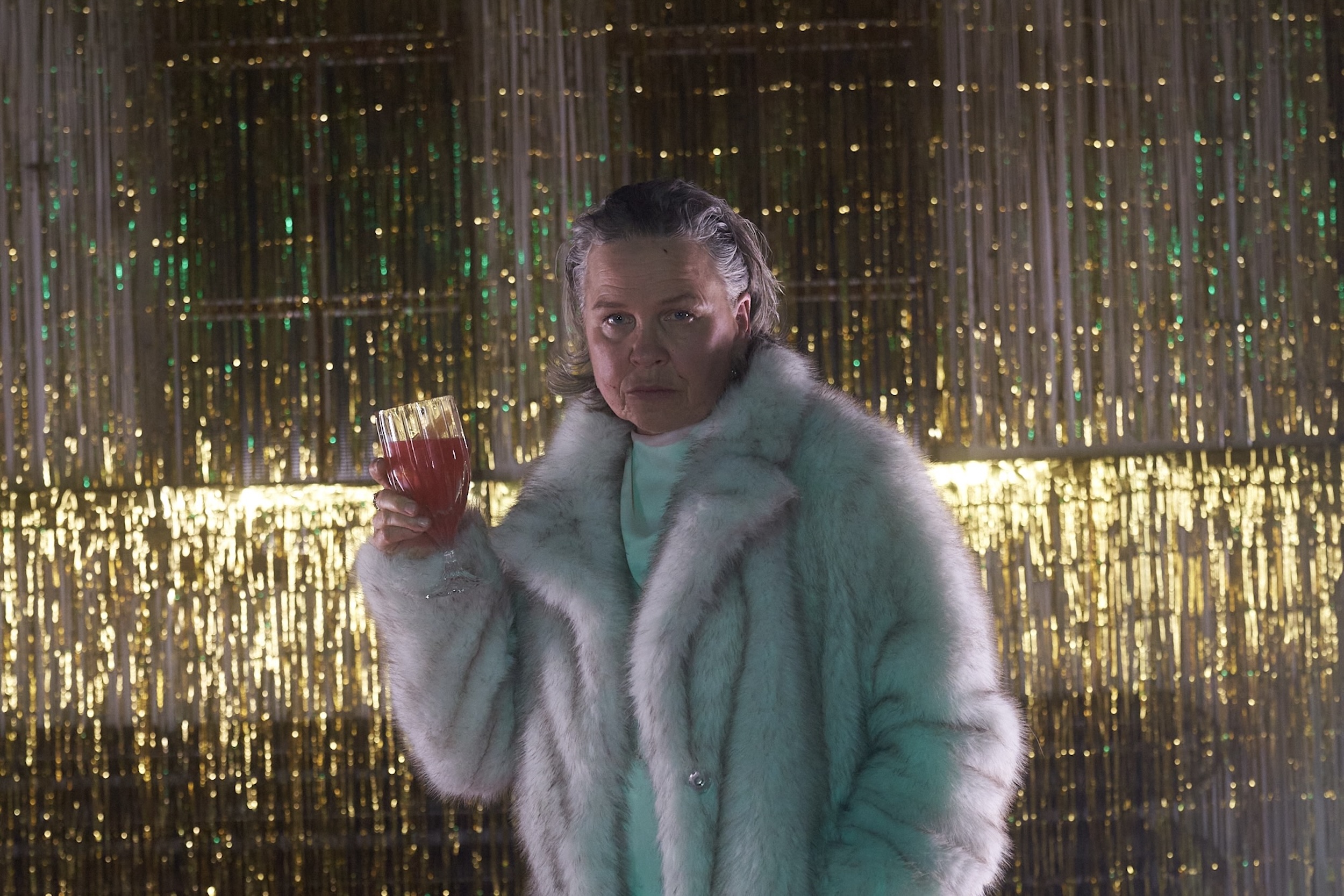 Person in hellem Kunstpelzmantel hält ein Glas mit rotem Getränk in der Hand, steht vor golden glitzerndem Vorhang in bühnenartiger Atmosphäre; das Gesicht ist unkenntlich gemacht.