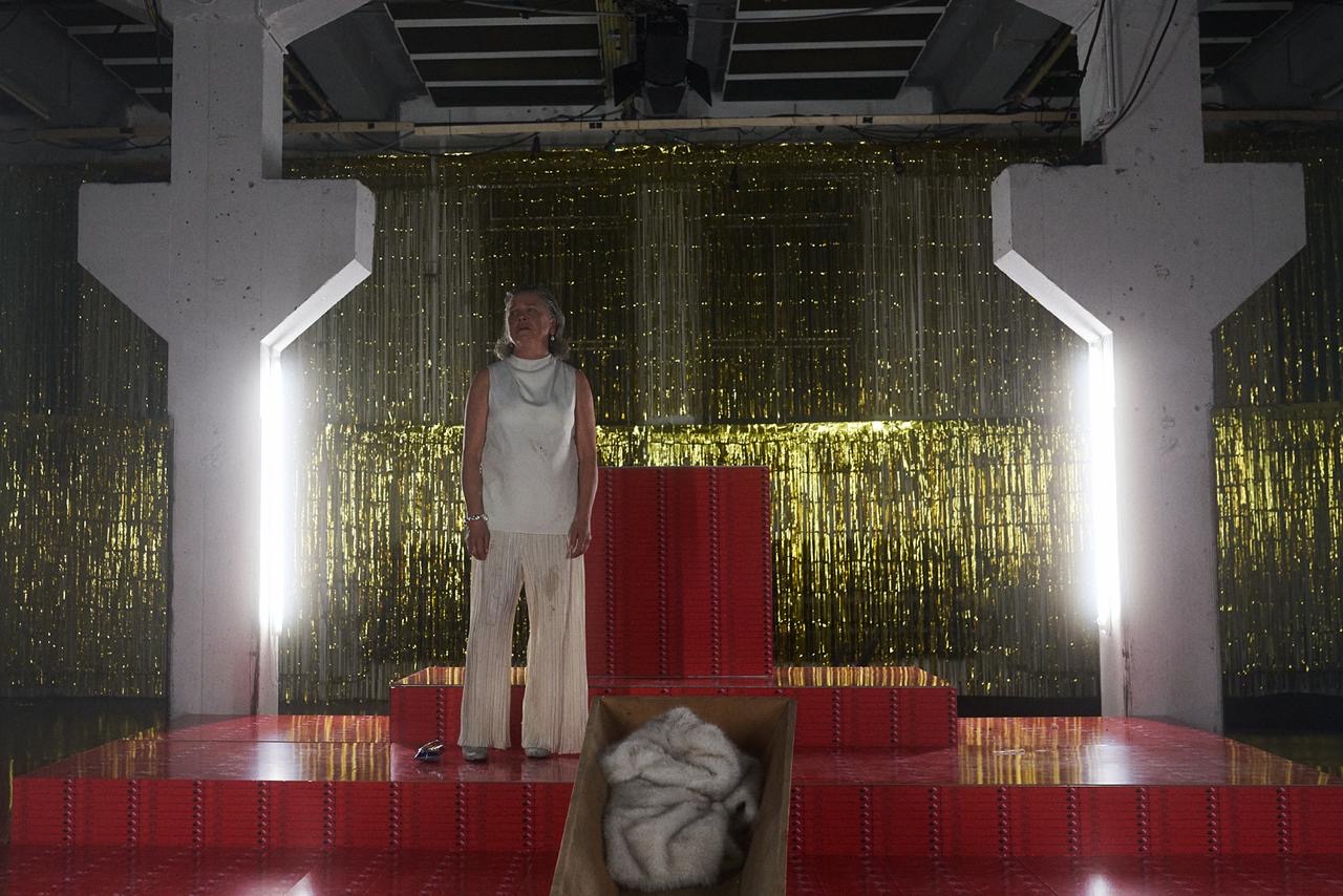 Person in heller, ärmelloser Oberbekleidung und weiter Hose steht auf einer roten, stufigen Bühnenplattform; im Vordergrund ein offener Holzkasten mit heller Felldecke, im Hintergrund goldener Glitzervorhang und massive Betonsäulen eines Industrie- oder Theaterraums.