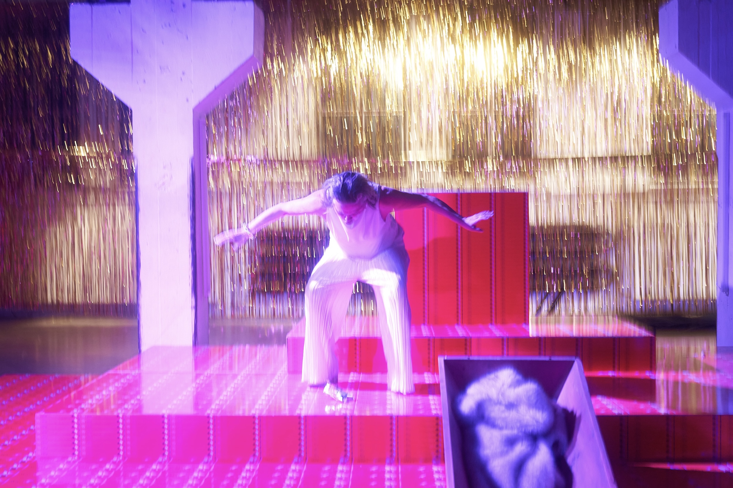Person in weißem, ärmellosem Oberteil und weiter Hose steht zentral auf einer treppenartigen, leuchtend pink‑roten Bühnenplattform; im Vordergrund ein offener, gepolsterter Kasten, links und rechts stehen massive helle Säulen vor schwarzem Hintergrund.