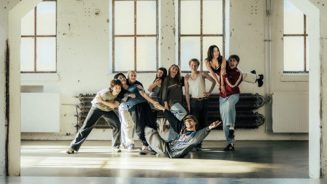 Eine Gruppe von Menschen posiert in einer industriellen Umgebung mit großen Fenstern. Einige stehen, andere sitzen oder lehnen sich aneinander, während eine Person hochgehoben wird.