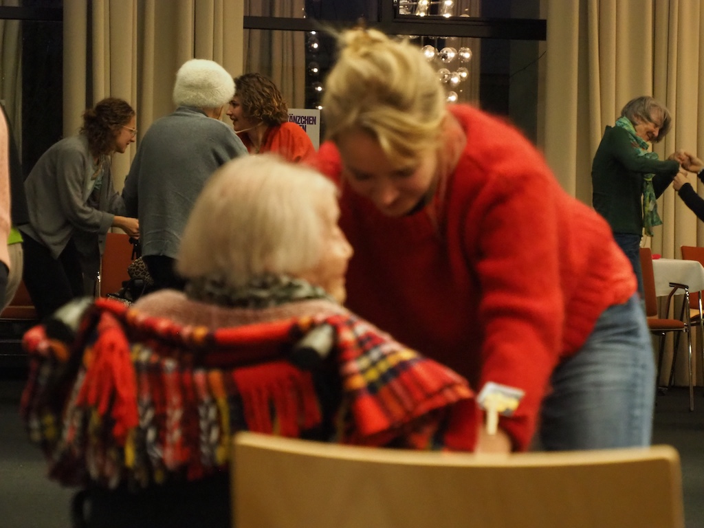 Eine intime Interaktion zwischen mehreren Personen in einem Veranstaltungsraum. Im Vordergrund ist eine Person in einem roten Pullover zu sehen, die sich zu einer anderen Person hinunterbeugt, die eine farbenfrohe Decke trägt. Im Hintergrund sind weitere Personen in Gesprächen vertieft. Die Szene vermittelt eine warme, fürsorgliche Atmosphäre.