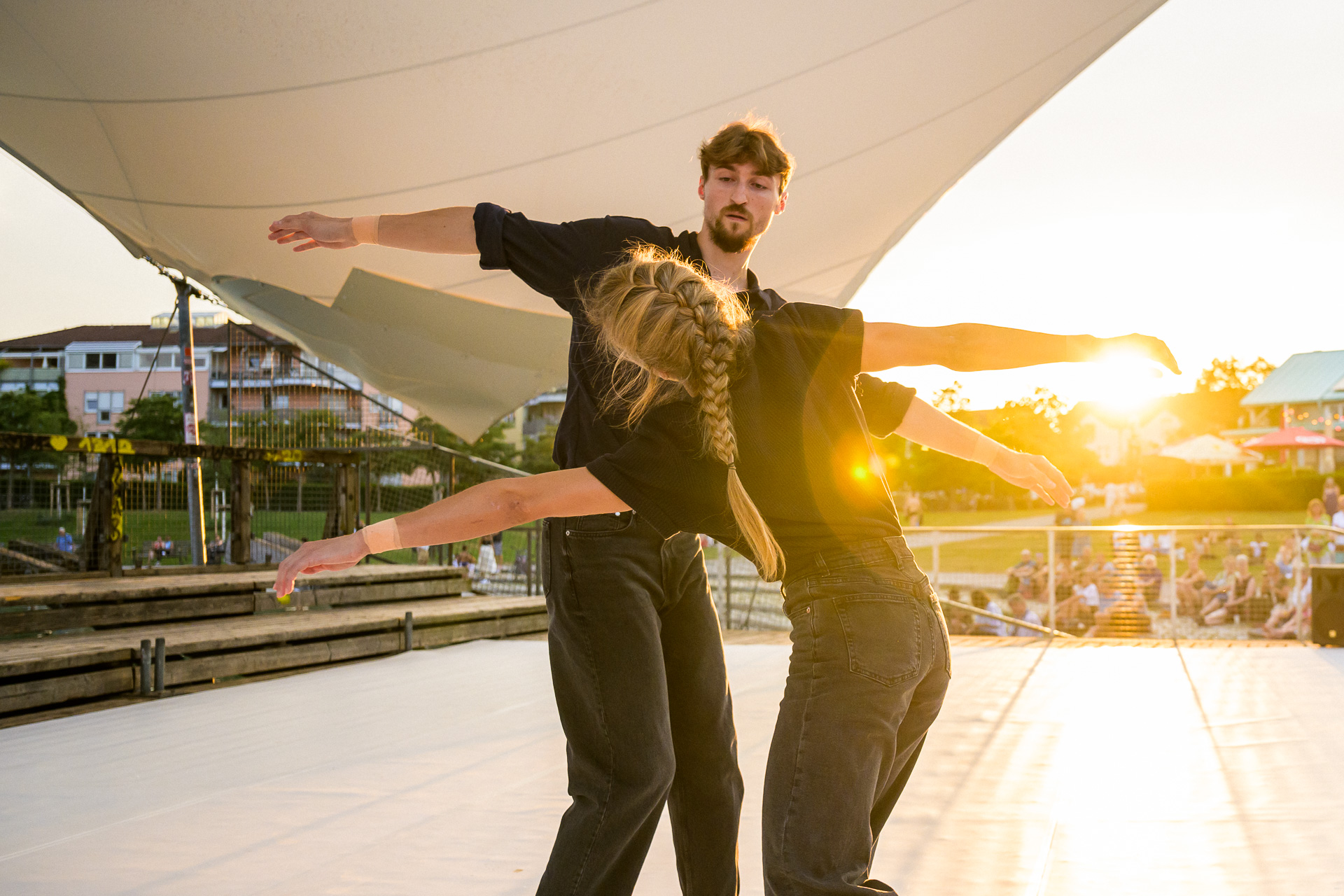 Zwei Personen tanzen auf einer Bühne im Freien bei Sonnenuntergang. Die Frau hat blondes Haar, das zu einem Zopf geflochten ist, und lehnt sich mit ausgestreckten Armen nach hinten. Der Mann trägt einen Bart und schaut konzentriert auf sie. Beide tragen schwarze Kleidung, und im Hintergrund sind Zuschauer sowie Gebäude zu sehen. Die Szene ist in warmes Sonnenlicht getaucht.  Quellen [1] TT-c-Doradzillo.jpg https://pplx-res.cloudinary.com/image/upload/v1739445568/user_uploads/OLfpUDeiyrooqeN/TT-c-Doradzillo.jpg