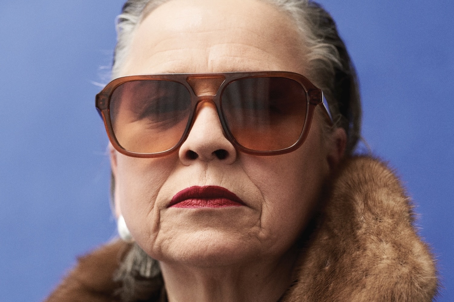 Ein professionelles Porträtfoto einer Person mit schulterlangem, zurückgekämmtem grauem Haar, die einen schwarzen Rollkragenpullover trägt. Das Bild zeigt einen intensiven, direkten Blick mit ausdrucksstarken hellblauen Augen. Der Gesichtsausdruck ist nachdenklich und ernst. Das Foto ist vor einem dunklen, unscharfen Hintergrund aufgenommen und zeichnet sich durch eine hochwertige Beleuchtung aus, die die Gesichtszüge der Person betont. Die Aufnahme vermittelt eine starke Präsenz und emotionale Tiefe.