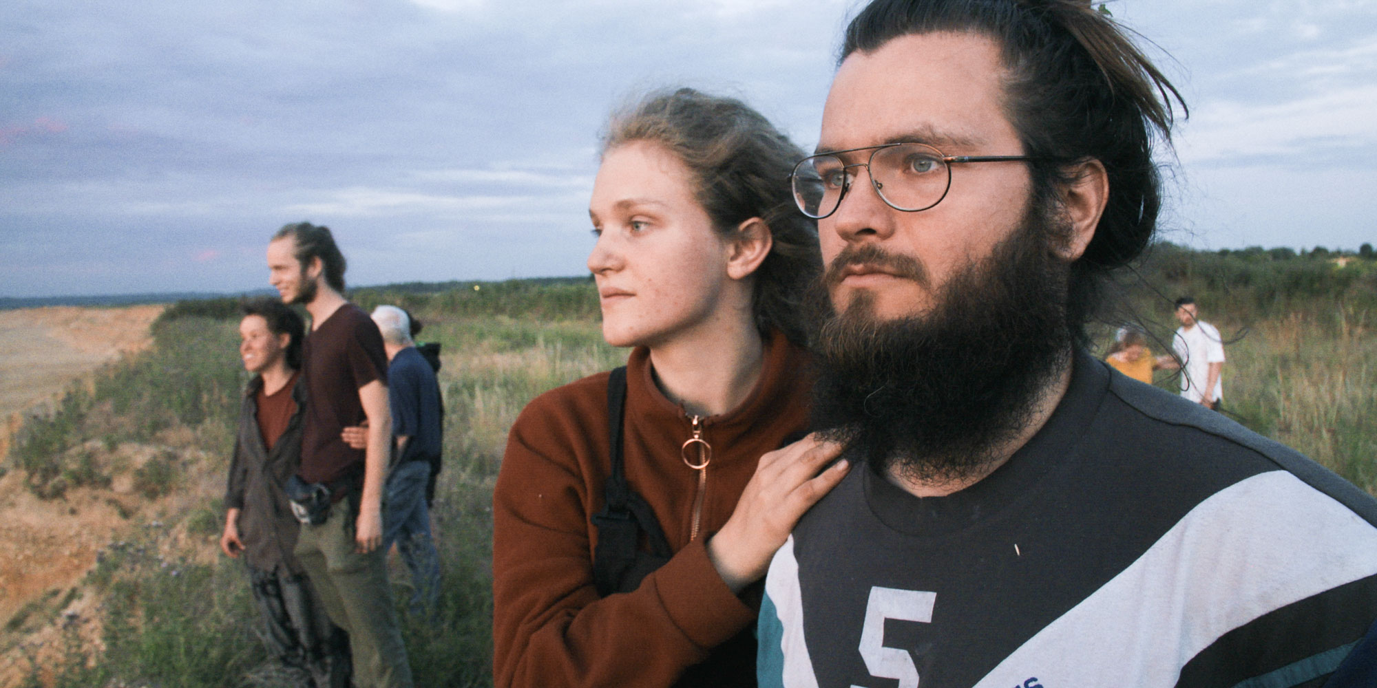 Eine Gruppe junger Menschen steht auf einem kargen Feld bei bewölktem Himmel. Im Vordergrund sind zwei Personen zu sehen - eine mit langen Haaren in braunem Pullover und eine mit Brille und langem Bart in einem Sweatshirt mit der Zahl 5. Die Atmosphäre wirkt entspannt und nachdenklich