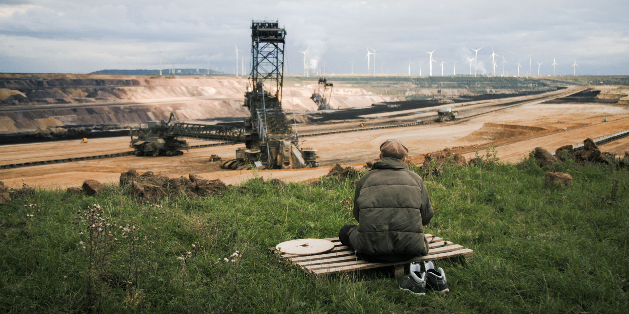 Eine Person in einer dicken Winterjacke sitzt auf einer Bank oder Palette und blickt auf einen großen Tagebau. Im Hintergrund sind massive Bergbaumaschinen zu sehen, darunter ein großer Schaufelradbagger. Am Horizont stehen mehrere Windkraftanlagen. Die Szene zeigt den Kontrast zwischen fossiler und erneuerbarer Energiegewinnung.