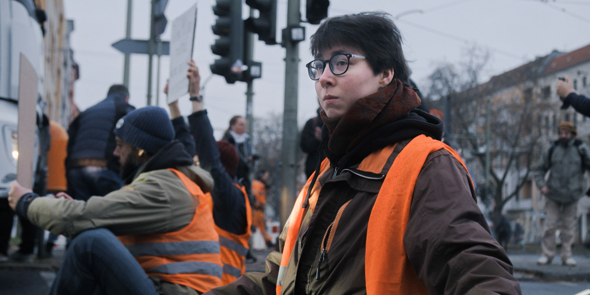 Eine Person mit Brille, kurzen dunklen Haaren und mehreren Piercings trägt eine orangefarbene Warnweste über dunkler Winterkleidung. Im Hintergrund sind weitere Menschen mit Warnwesten zu sehen, die Schilder hochhalten, was auf eine Demonstration oder Protestaktion hindeutet. Die Szene spielt in einem urbanen Umfeld mit Ampeln und Gebäuden im Hintergrund.