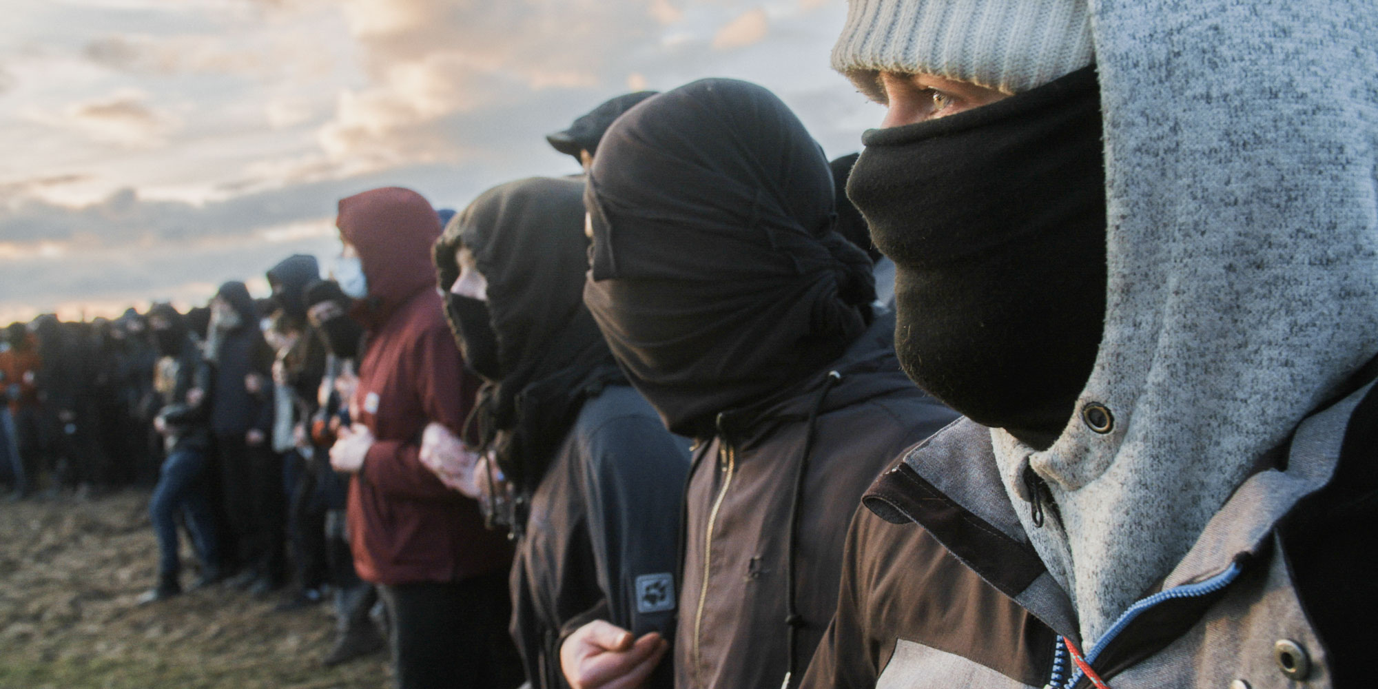 Eine Reihe von Menschen steht in der Dämmerung auf einem Feld. Sie tragen dunkle Kleidung und haben ihre Gesichter mit Tüchern und Masken vermummt. Die Stimmung wirkt geheimnisvoll und organisiert.