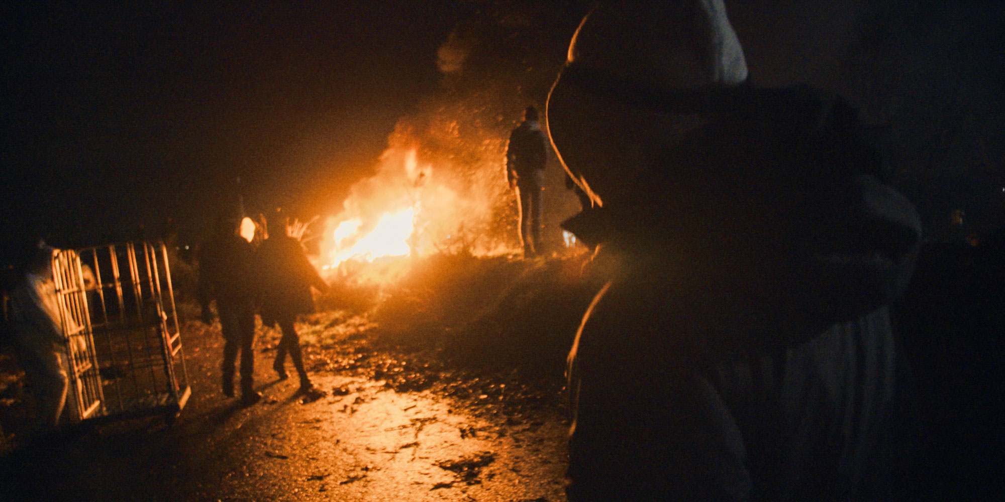 Eine nächtliche Szene mit einem großen Feuer, das orange Licht auf die Umgebung wirft. Im Vordergrund sind Silhouetten mehrerer Personen zu erkennen, sowie ein metallenes Gitter oder Zaunteil. Die Szene strahlt eine intensive, fast mystische Atmosphäre aus.