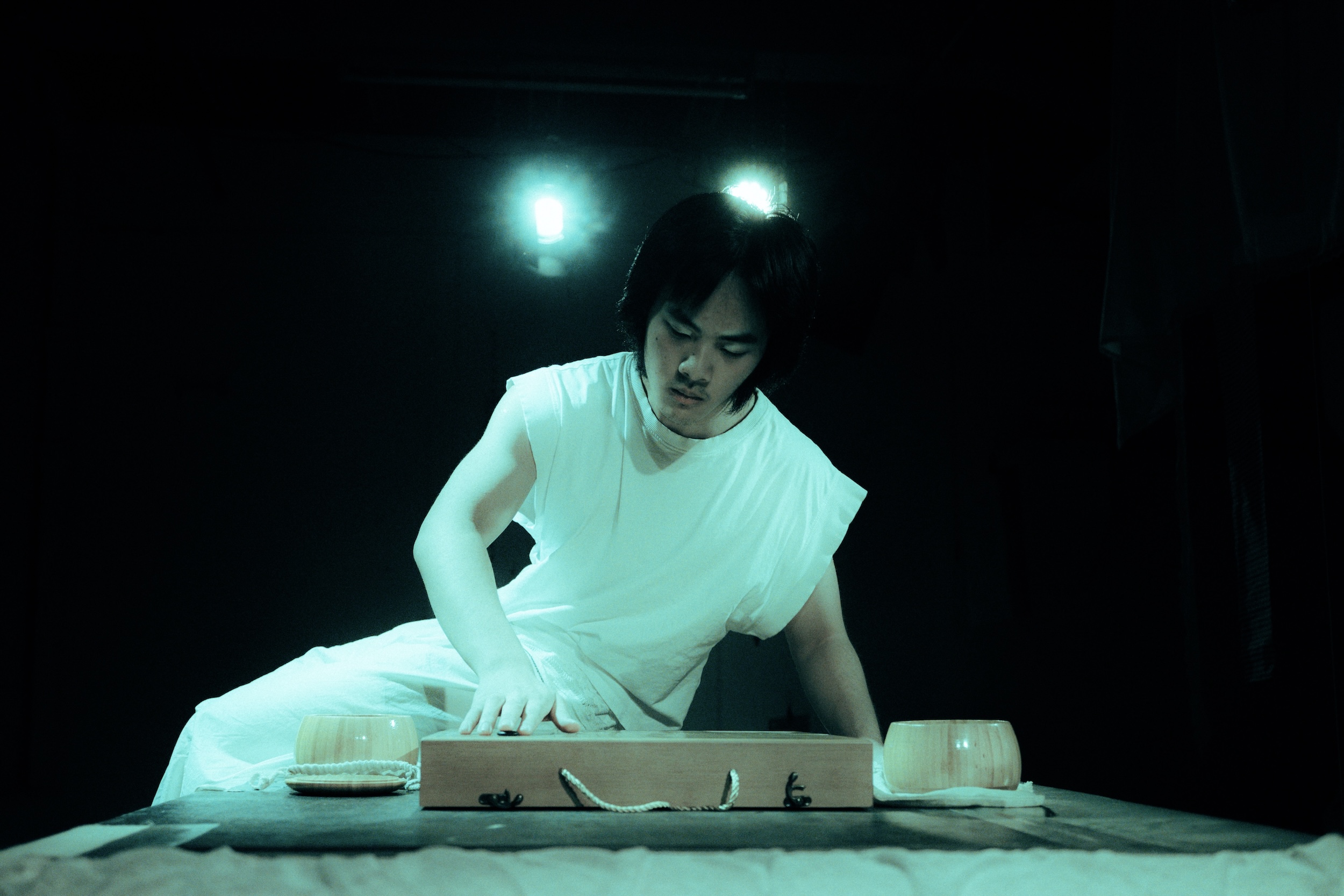 Eine einzelne Person im weißen Gewand sitzt an einem niedrigen Holztisch und blickt konzentriert nach unten, zwei Schalen stehen davor