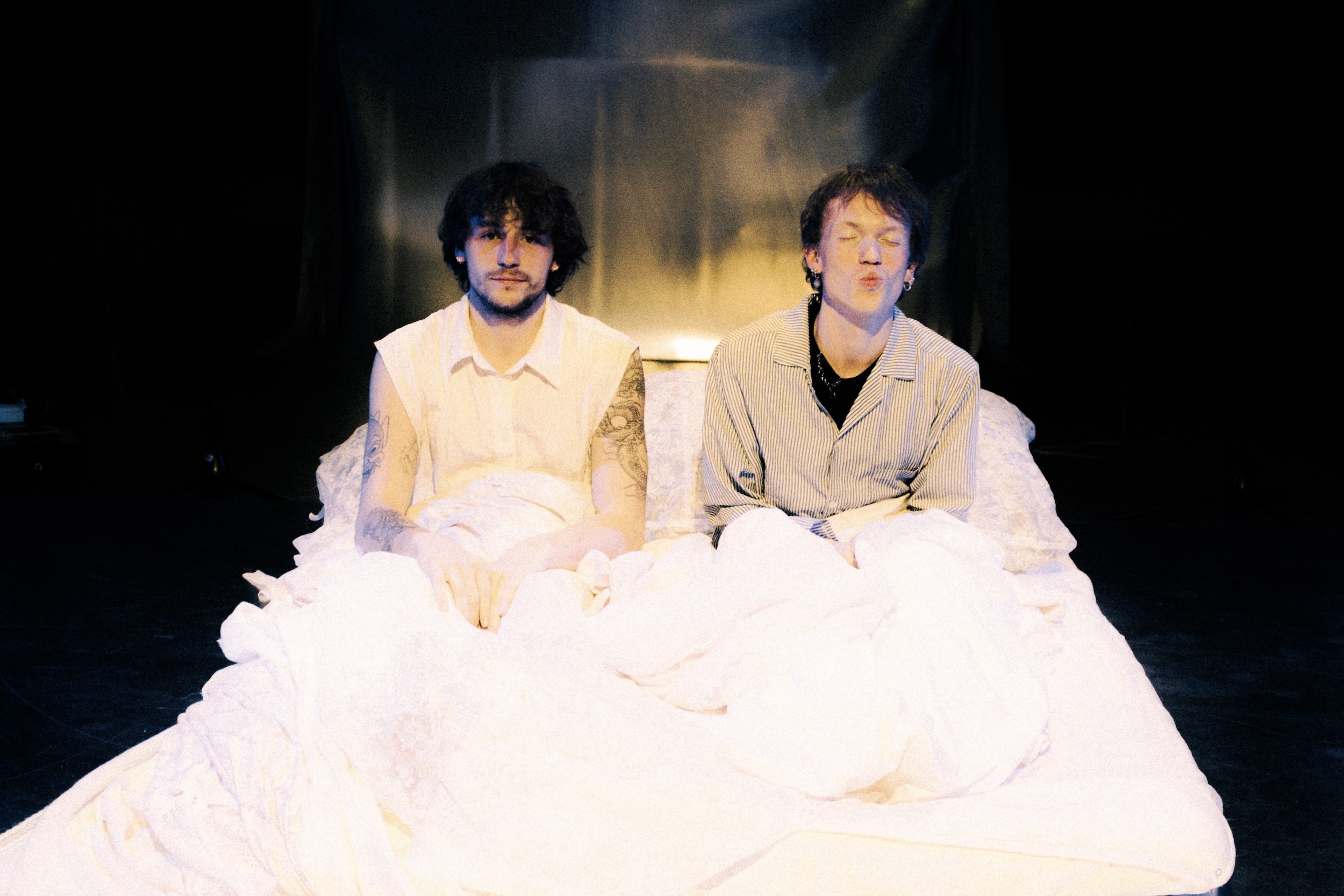 Zwei Personen sitzen nebeneinander auf einem hellen Bett, in weiße Stoffe gehüllt und von einer warmen Bühne beleuchtet