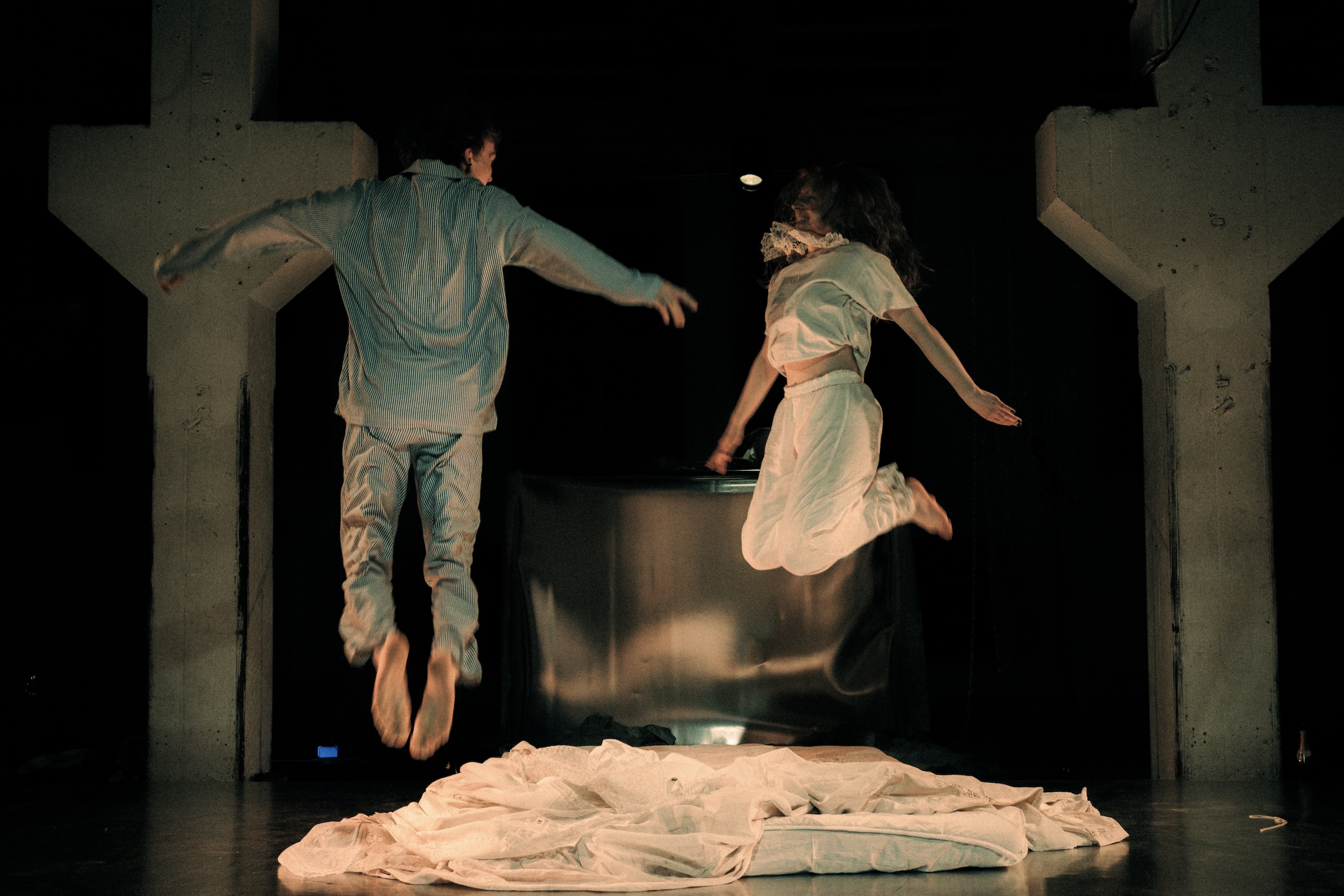 Zwei Performer*innen springen gleichzeitig in die Luft, beide ganz in Weiß gekleidet, im Hintergrund stehen große Säulen