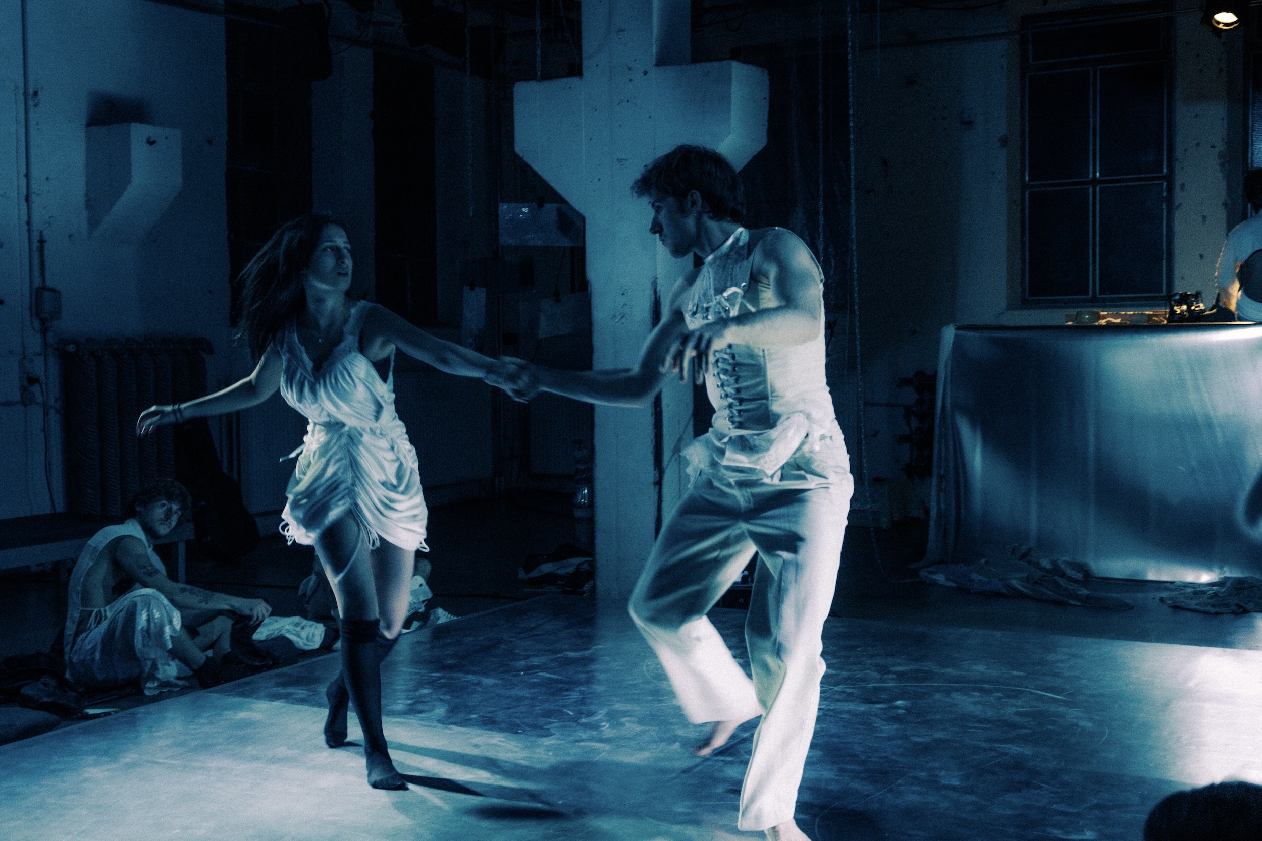 Zwei Tänzer*innen in weißen Outfits halten sich an den Händen und tanzen, im Hintergrund sind weitere Personen und ein weiß gedeckter Tisch zu sehen.