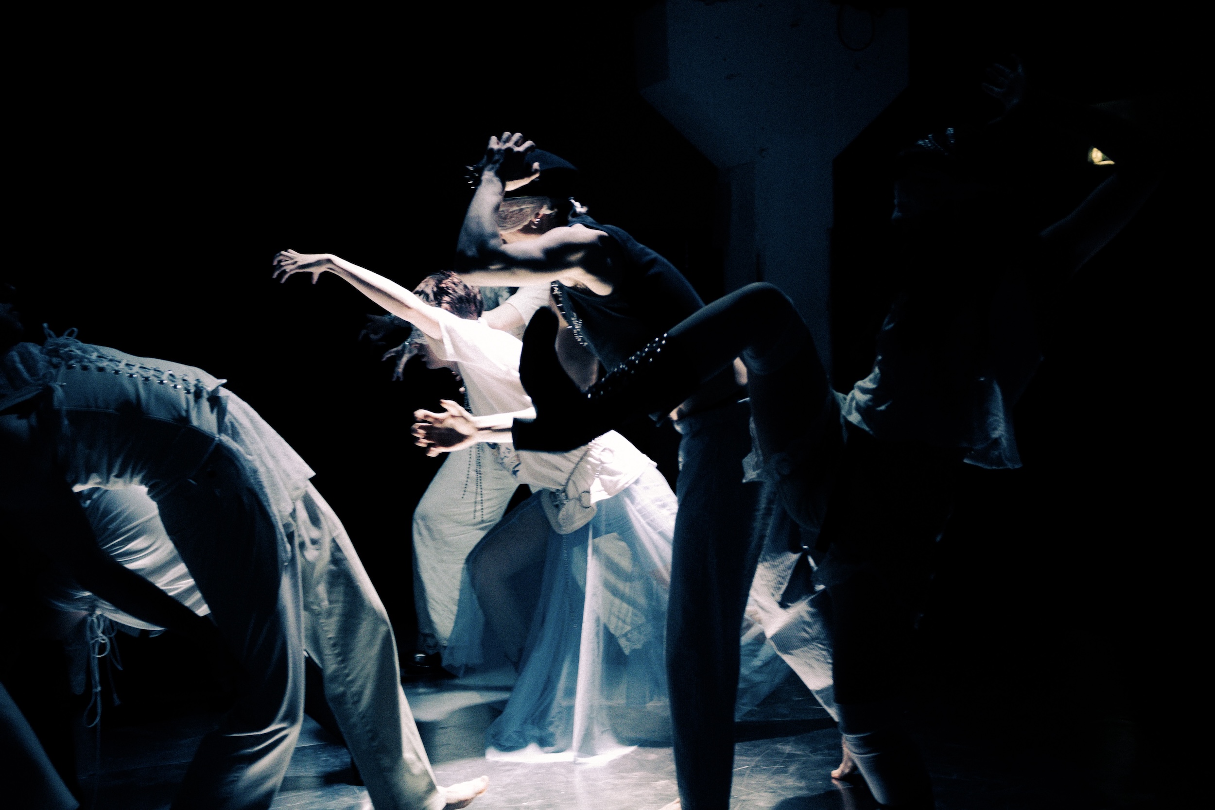 Eine Gruppe von Performer*innen in weißen und schwarzen Kostümen tanzt auf einer dunklen Bühne, dynamisch ausgeleuchtet und in ausdrucksvollen Körperhaltungen.