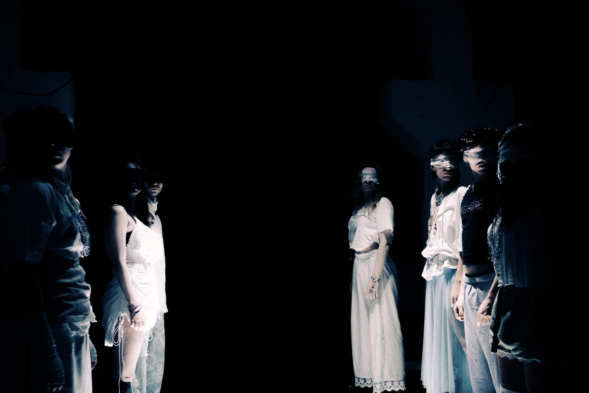 Zwei Gruppen stehen sich gegenüber in einer dunklen Bühnenatmosphäre, die meisten Personen tragen helle Kleidung und blicken nach vorne.