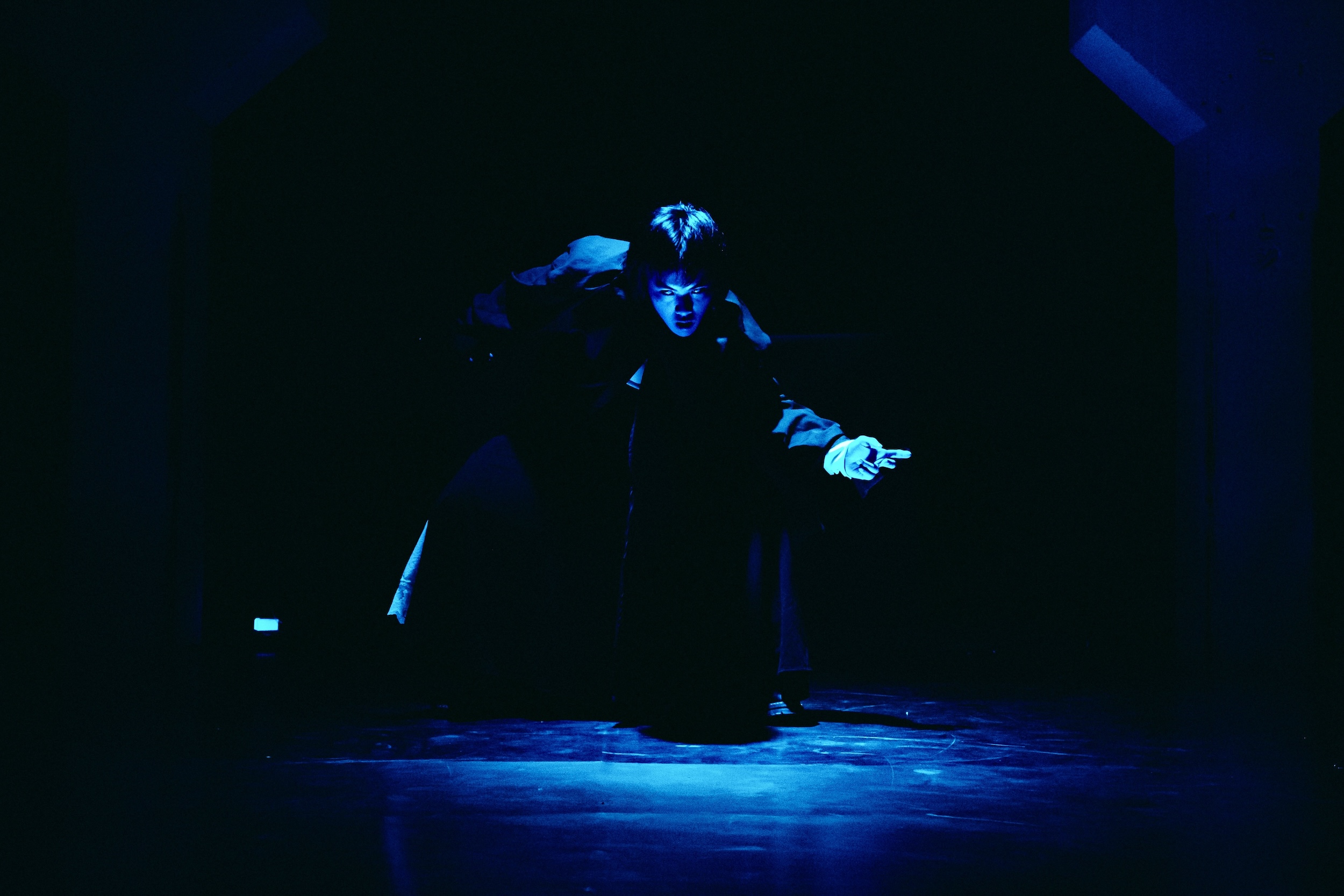 Eine dramatisch beleuchtete Person in dunkler Kleidung steht auf einer Bühne mit kräftigem blauem Licht und expressiver Gestik.
