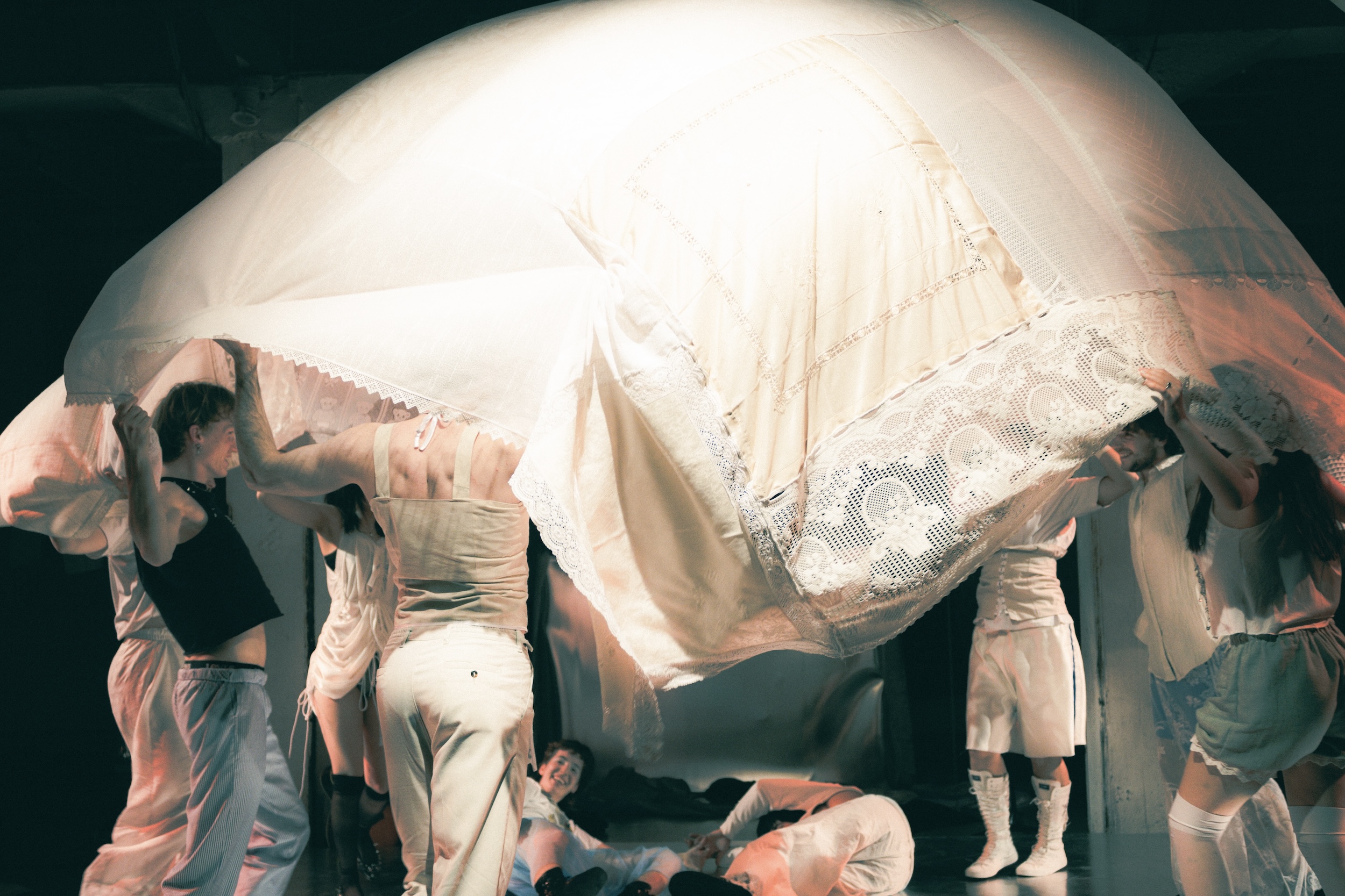 Eine Gruppe junger Menschen sitzt und liegt entspannt auf einem weißen Bühnenboden, während jemand Gitarre spielt und aus einem Gefäß Dampf austritt