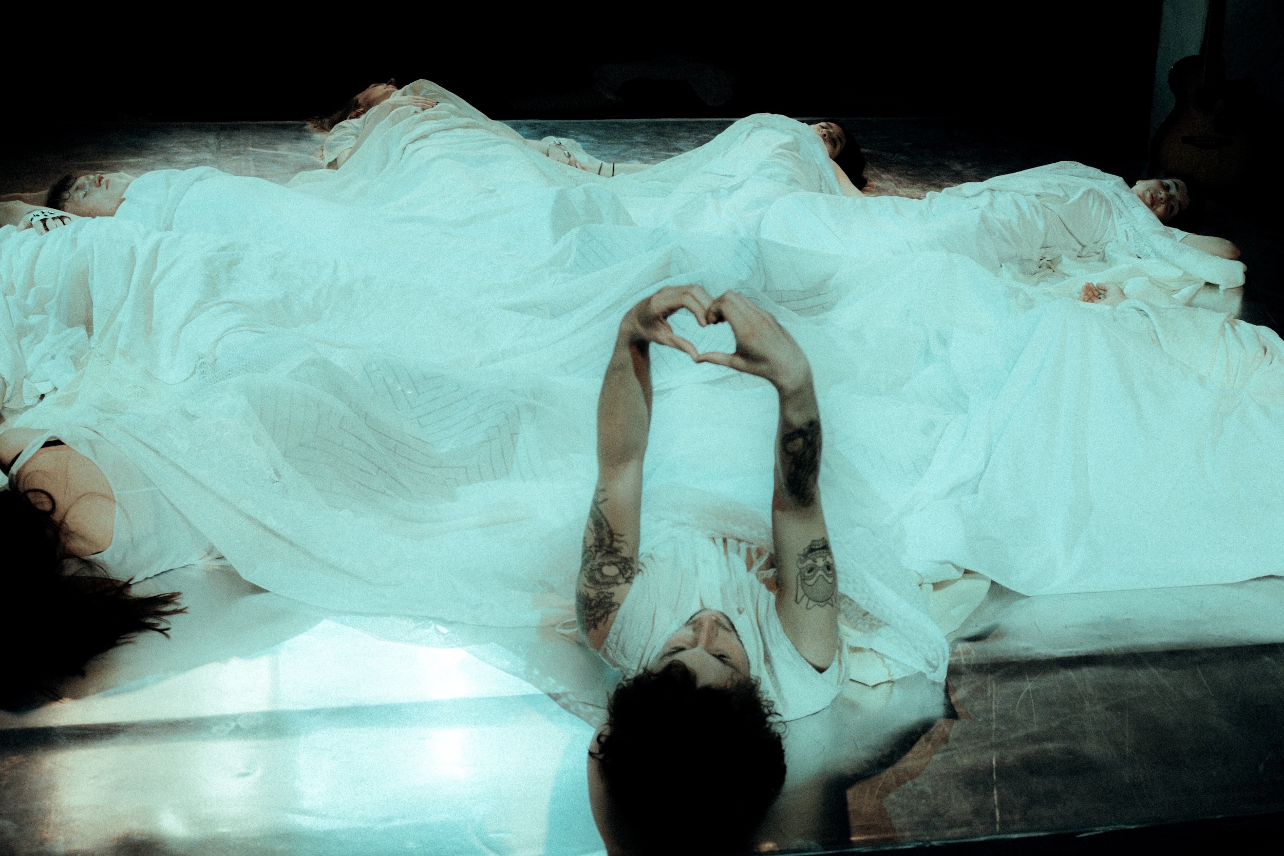 Mehrere Personen liegen nebeneinander unter weißen Decken auf einer Bühne, ein Mensch im Vordergrund formt mit den Händen ein Herzsymbol und blickt nach oben