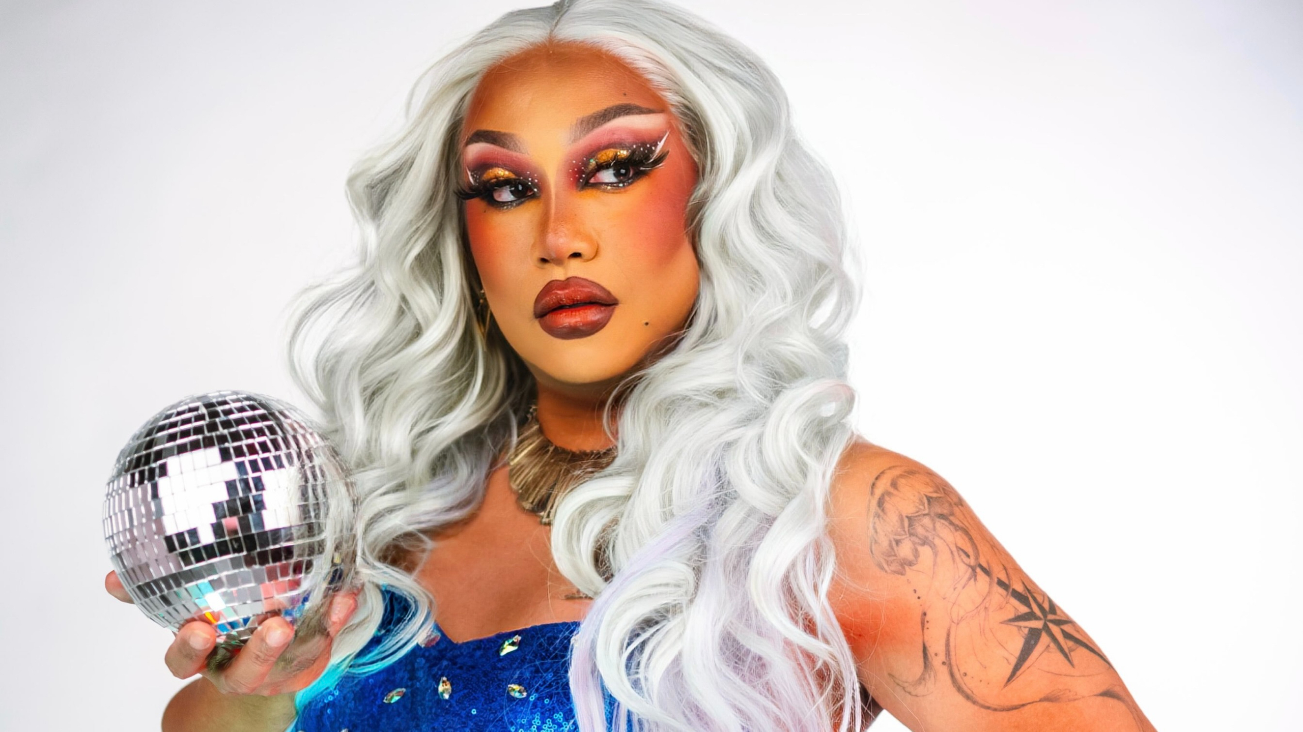 Eine Dragqueen mit langen, lockigen, weiß-grauen Haaren in glitzerndem blauen Kleid, hält eine silberne Diskokugel und trägt auffälligen Armschmuck sowie ein Tattoo mit Kompass und Qualle auf dem Oberarm.