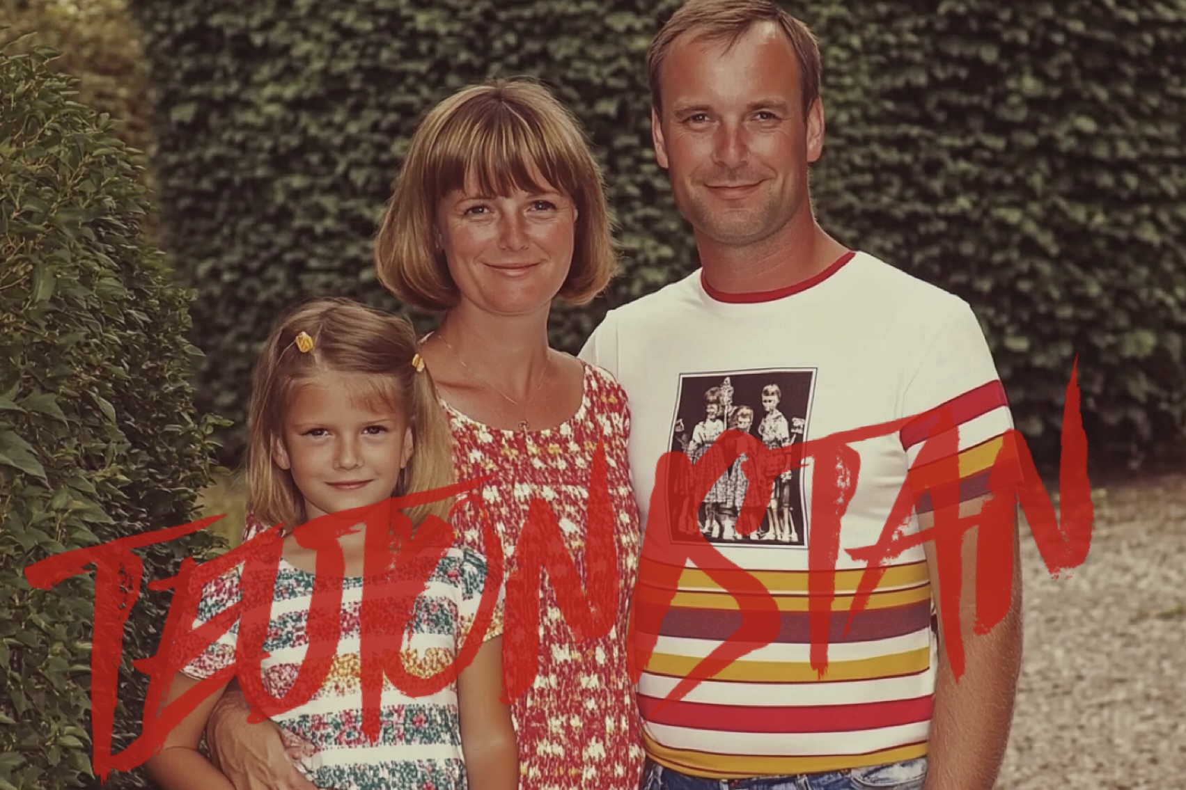 Eine weiße Mutter, ein Vater und ein Mädchen stehen eng beieinander im Freien vor grünen Hecken. Das Foto wirkt wie eine typische Familienaufnahme aus Deutschland aus den 1980ern oder 1990ern. Die Bekleidung ist sommerlich und bunt; der Mann trägt ein gestreiftes T-Shirt mit einem aufgedruckten Schwarzweiß-Foto von Kindern. Über das Bild ist in roter, handschriftlicher Schrift das Wort “TEUTONISTAN” geschrieben.
