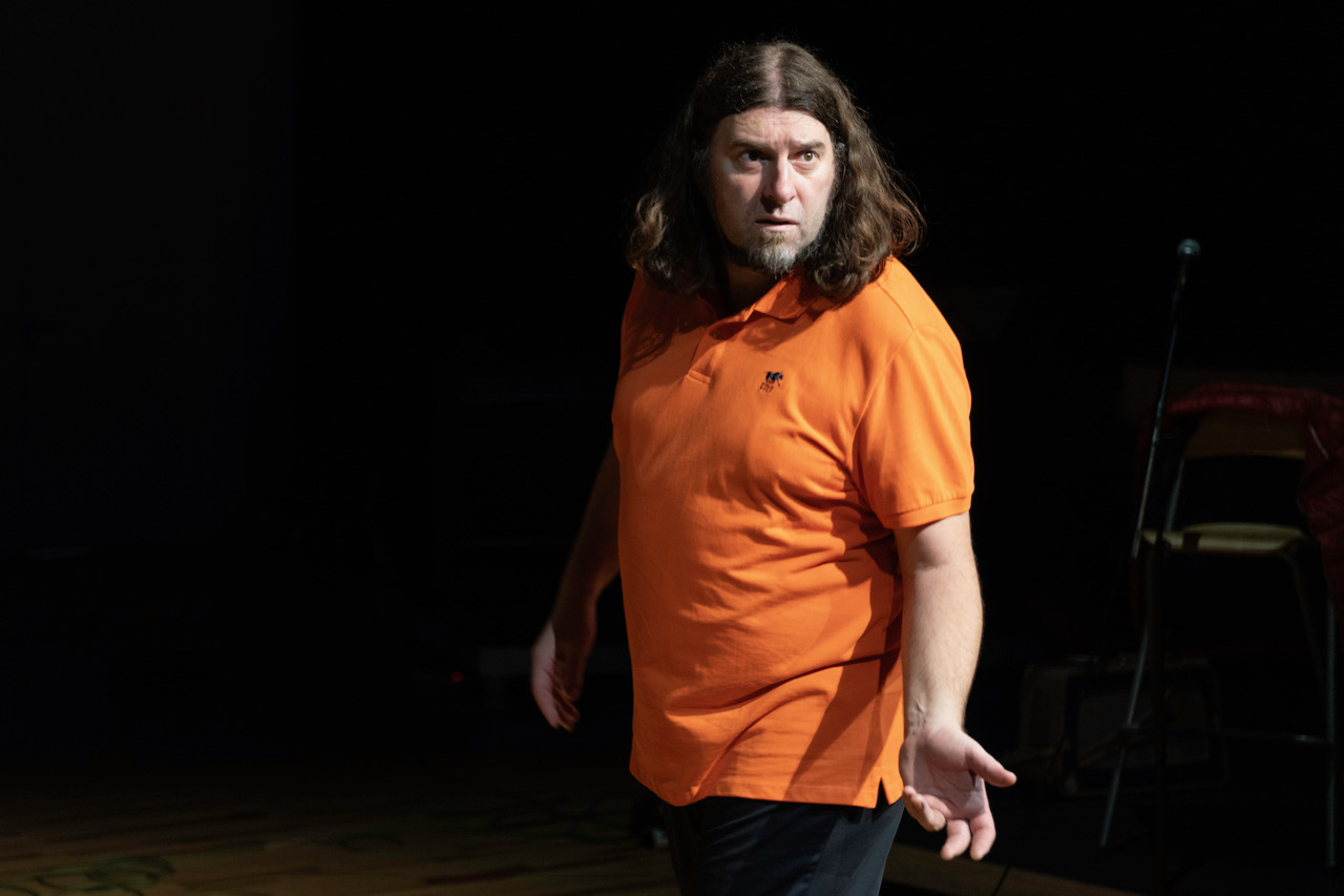 Person mit langen Haaren und orangem Poloshirt steht in einem abgedunkelten Bühnenraum, seitlich beleuchtet.