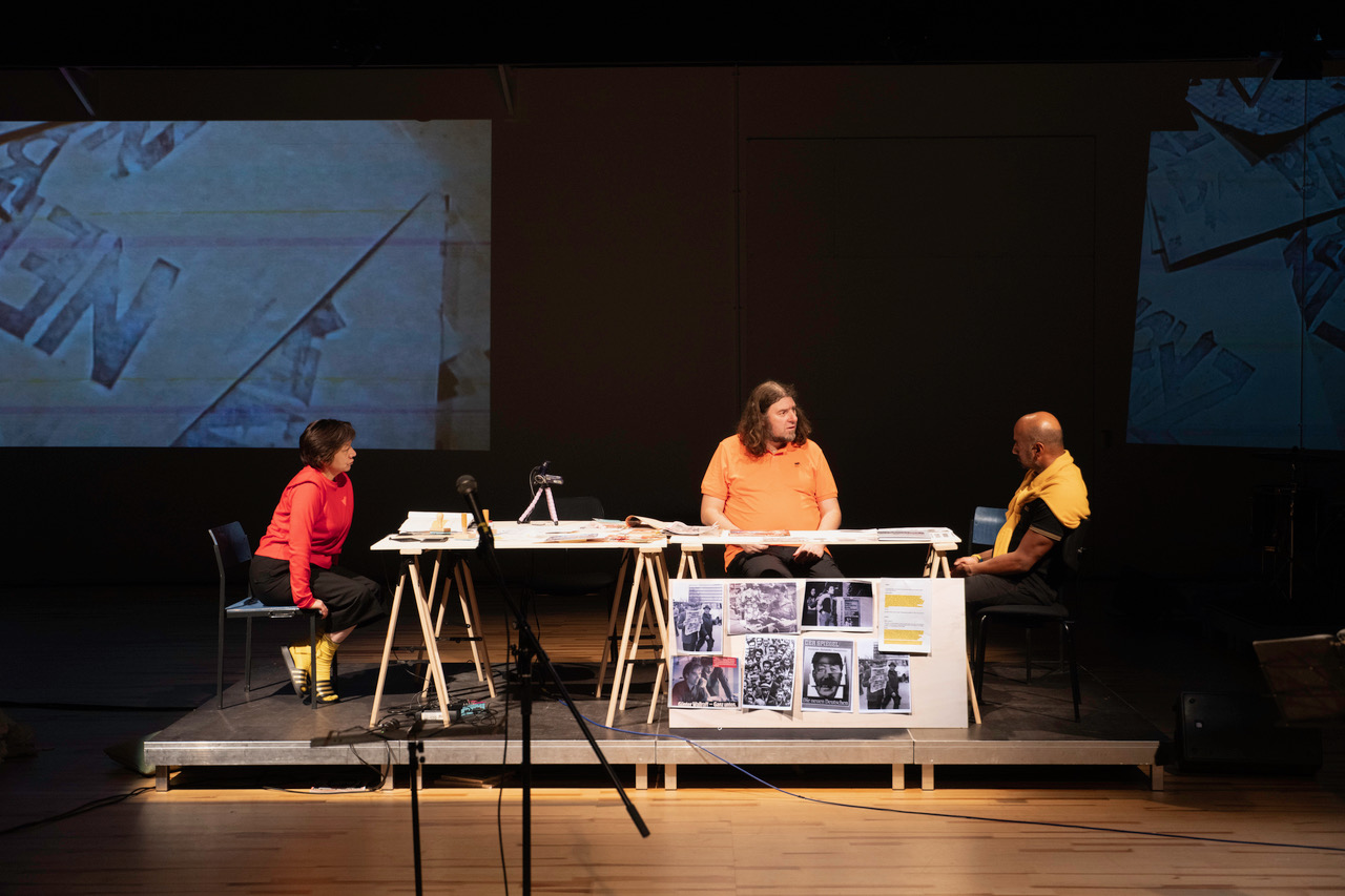 Drei Personen sitzen an einem langen Tisch auf einer Bühne und sind im Gespräch vertieft, die Wände im Hintergrund sind mit Doku-Material und Projektionen gestaltet.