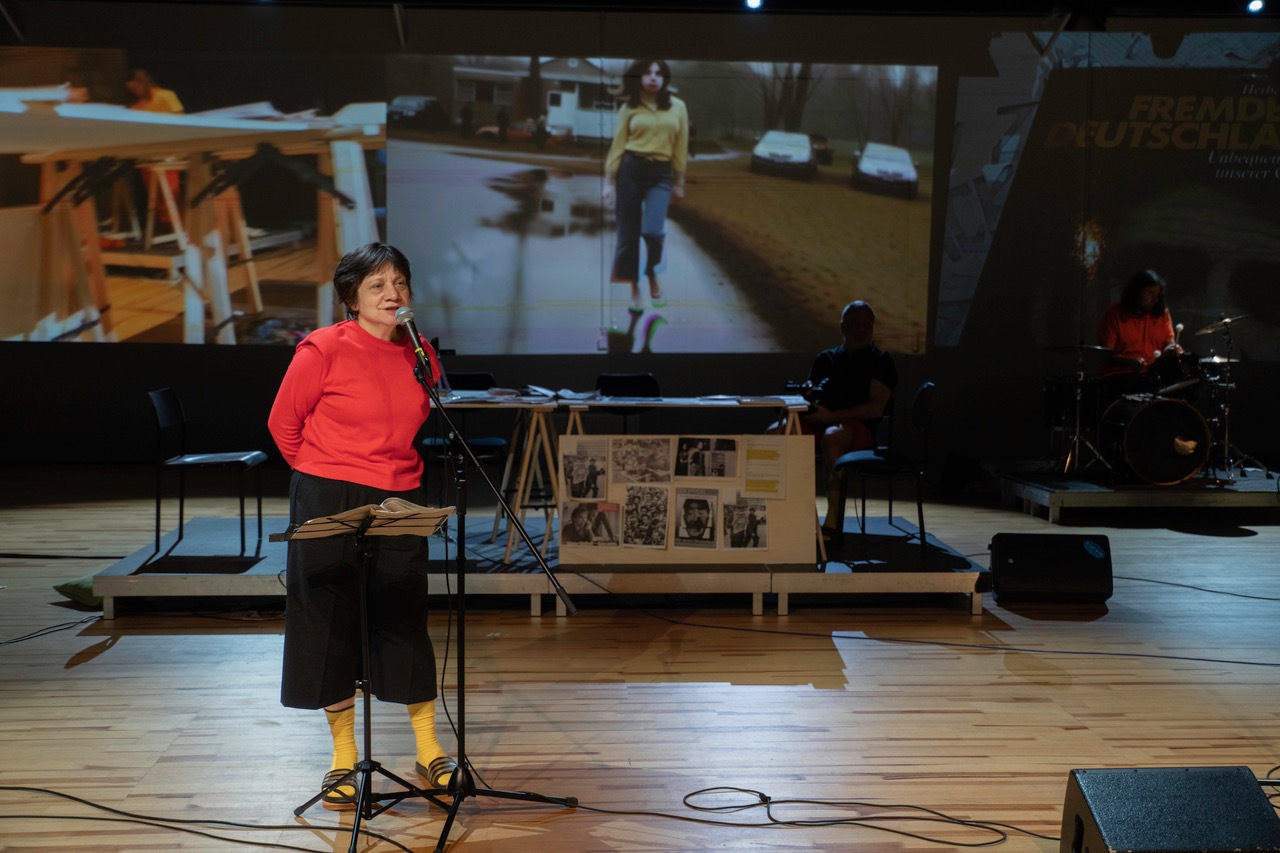 Frau mit rotem Shirt und gelben Socken steht an einem Mikrofon auf einer Theaterbühne, im Hintergrund eine große Videoprojektion und MusikerInnen.