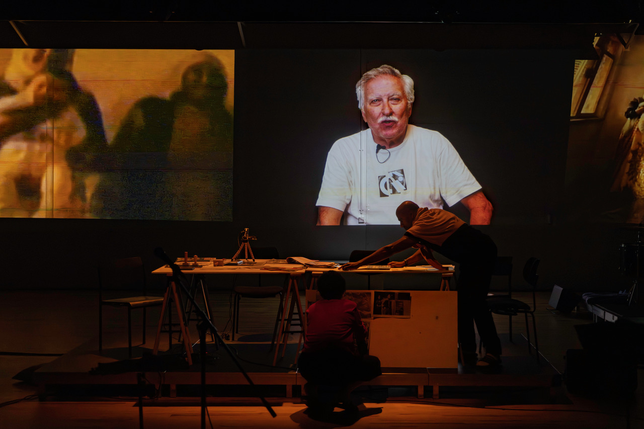 Große Videoprojektion zeigt eine Person im weißen T-Shirt; im Vordergrund sind zwei Menschen mit einer Arbeitsfläche voller Materialien bei einer Theaterperformance zu sehen