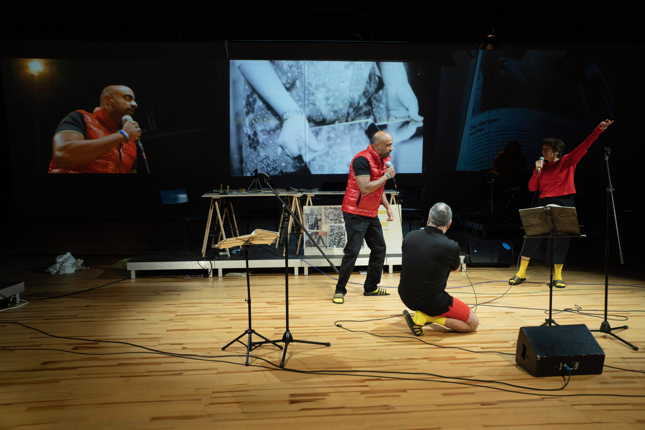 Drei Menschen auf der Bühne, zwei mit Mikrofon, einer kniet, alle wirken dynamisch in einer Performance, Projektionen und Materialien füllen den Bühnenhintergrund.