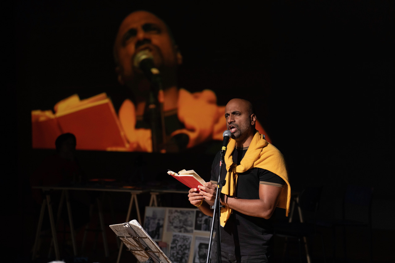 Ein Mann mit Buch und Mikrofon spricht auf der Bühne, trägt ein orangefarbenes Kleidungsstück über den Schultern. Im Hintergrund ist sein Gesicht groß projiziert und die Bühne mit Material sichtbar.