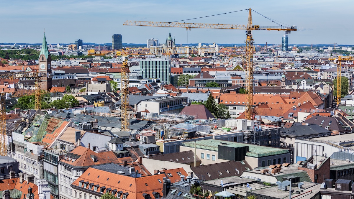 Blick über die Dächer von München mit zahlreichen Baukränen, roten Ziegeldächern und modernen Gebäuden. Im Vordergrund sind Bauarbeiten zu sehen, in der Ferne ragen Kirchtürme und Hochhäuser in den Himmel.