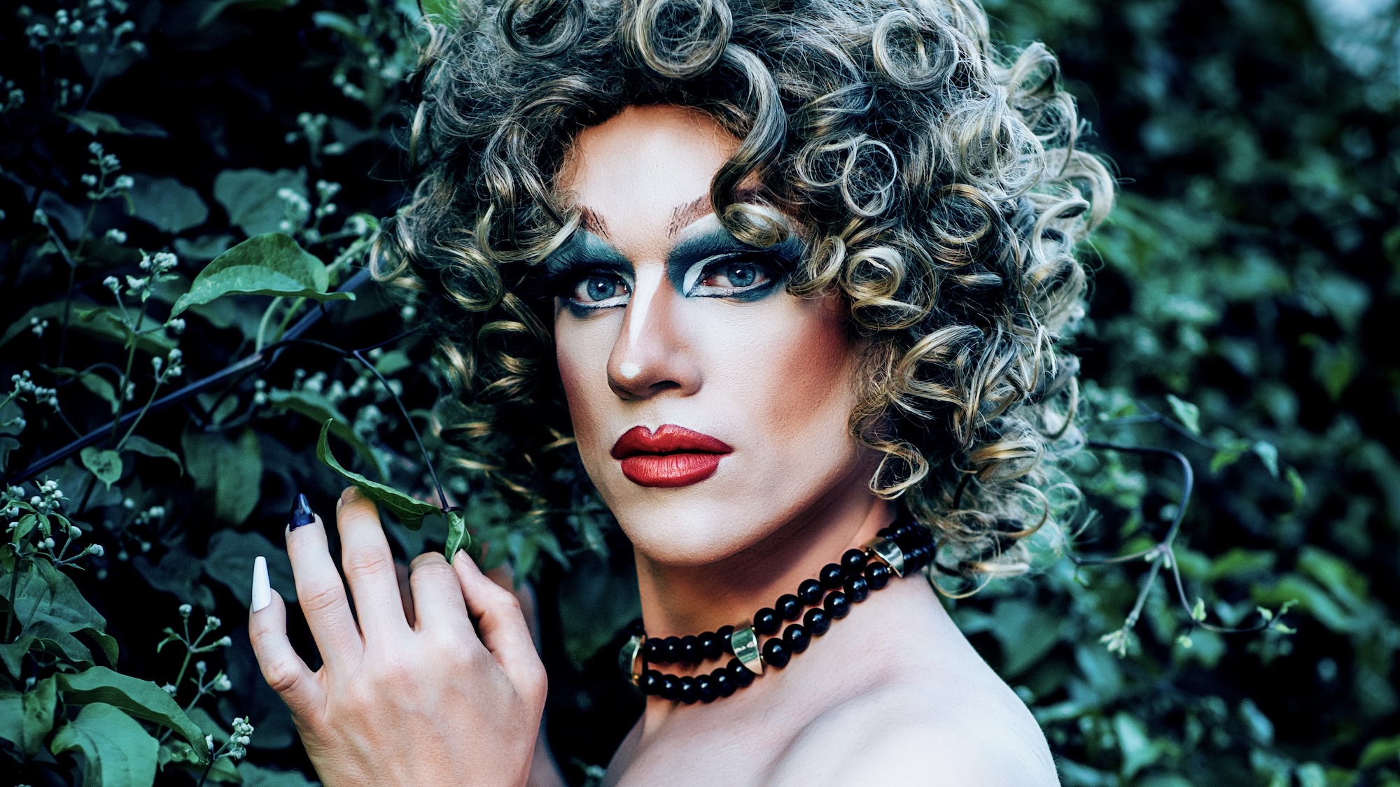 Eine Dragperson mit blonden, lockigen Haaren und einer Halskette aus schwarzen Perlen steht vor dichtem, grünem Blattwerk. Die Hand ist erhoben und berührt ein Blatt, die Nägel sind auffällig und farbig lackiert.