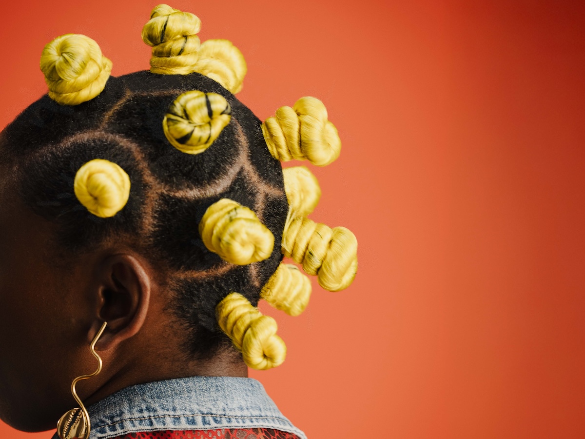 Der Kopf einer Person mit dunkler Haut und kunstvoll gestylten Haaren in Bantu-Knoten, die mit gelbem Garn umwickelt sind, ist vor einem roten Hintergrund zu sehen. Die Haarlinien sind präzise abgeteilt und auf der linken Seite trägt die Person einen auffälligen, goldfarbenen Ohrschmuck.