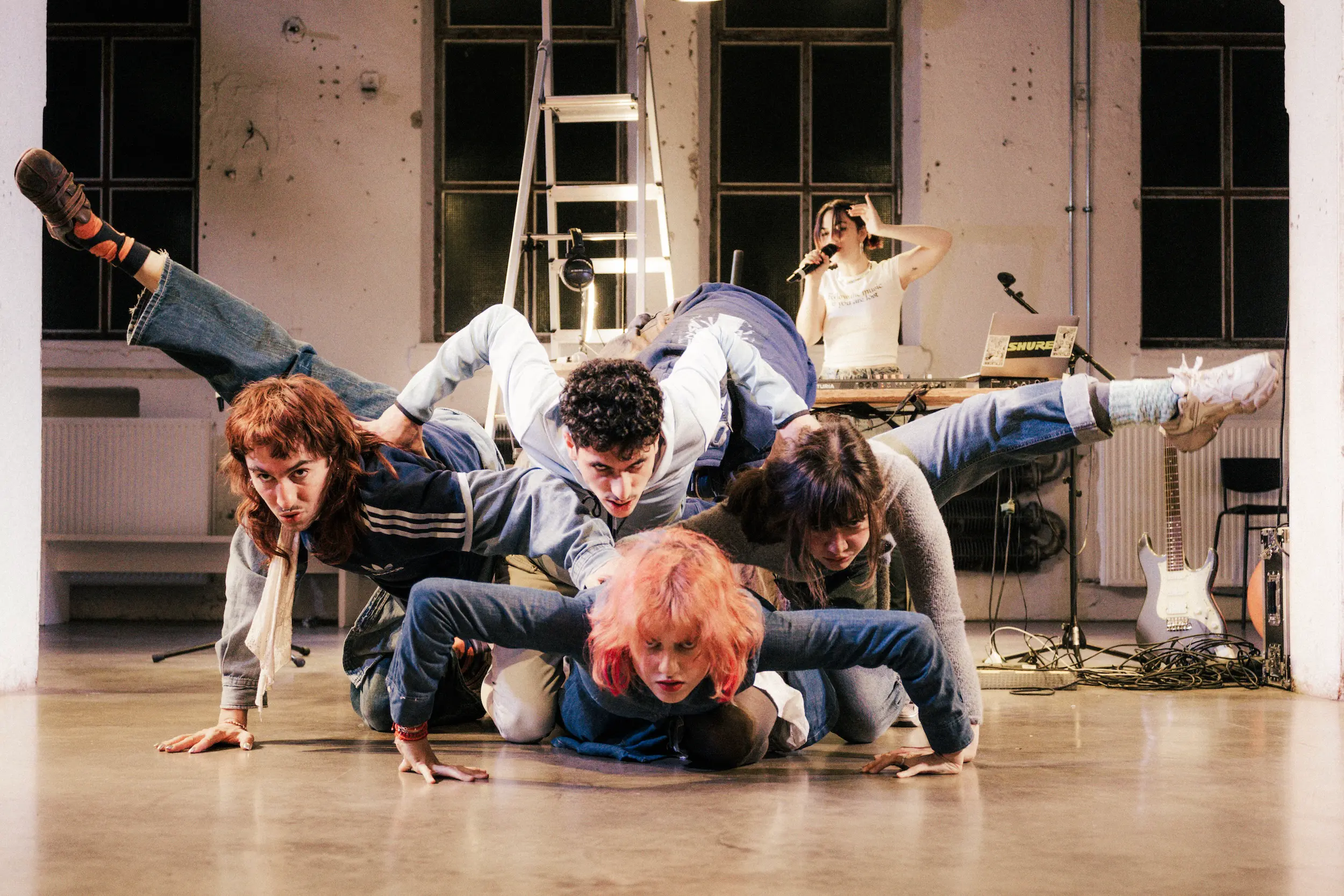 Farbige Bühnenszene in einem Industrie-Raum: Fünf Performende bilden eine dynamische, gestapelte Körperfigur auf dem Boden, während im Hintergrund eine Person an Laptop und Synthesizer steht und in ein Mikrofon singt.