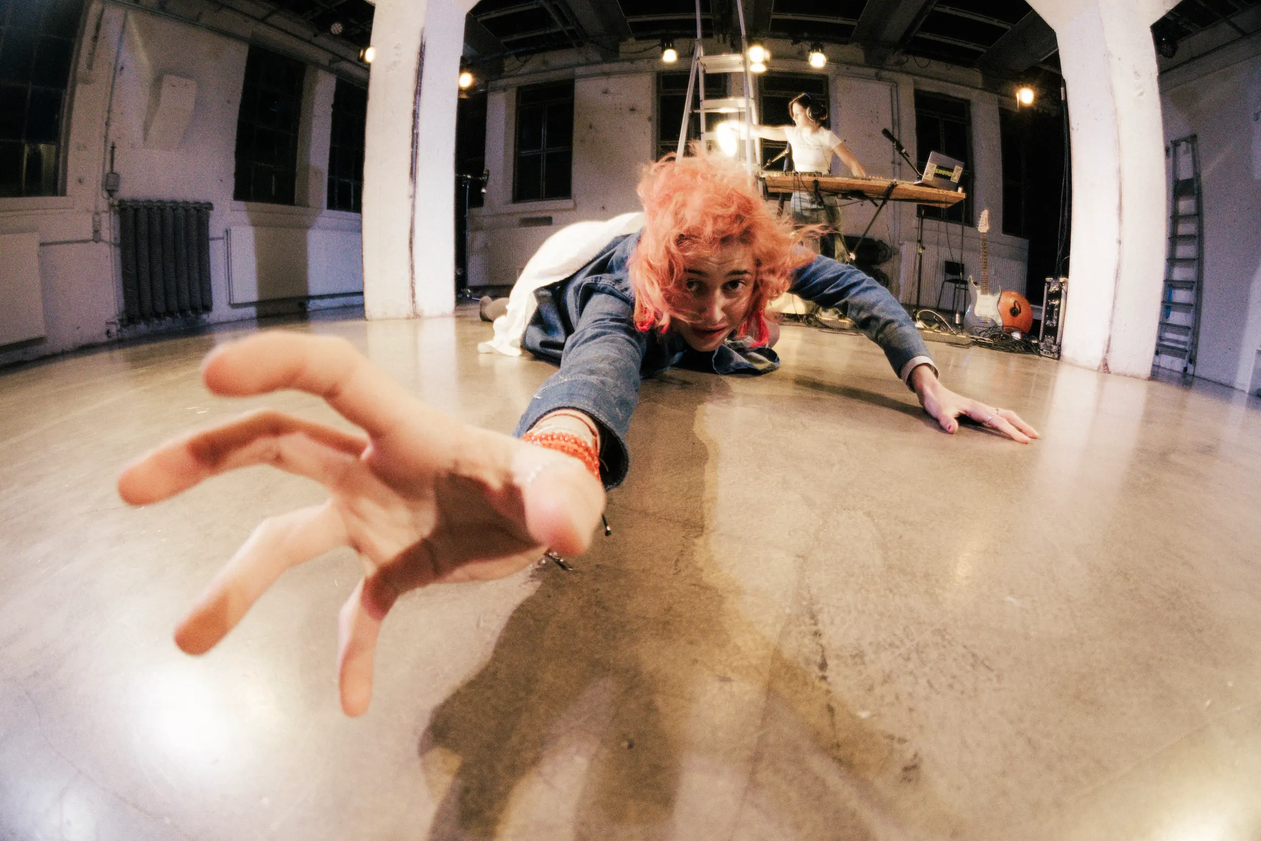 Eine Person mit gefärbten Haaren liegt bäuchlings auf dem Bühnenboden und streckt ihre Hand weit zur Kamera aus, im Hintergrund steht eine Musiker:in an einem Tisch mit Synthesizer und Mikrofon.