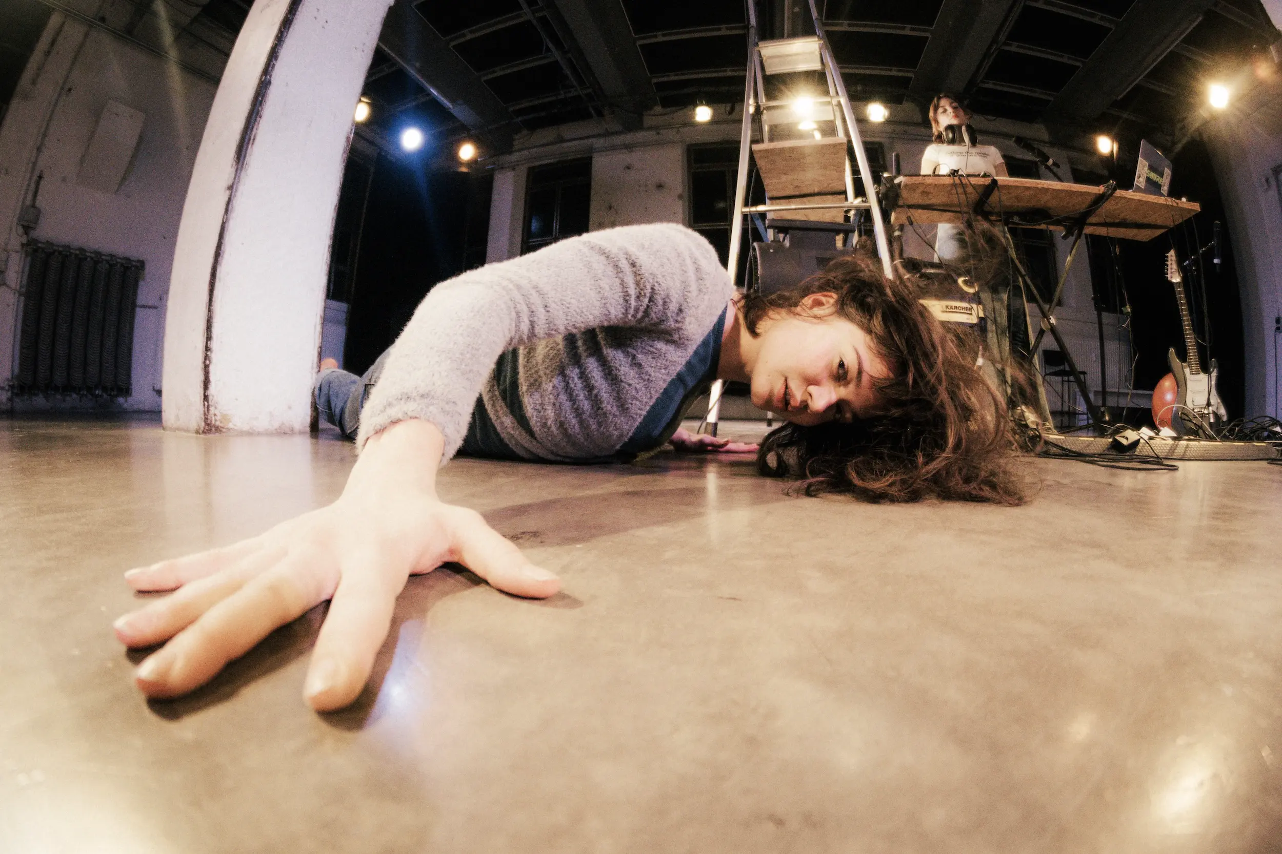 Eine Person liegt seitlich auf dem Bühnenboden und streckt die Hand zur Kamera, der Kopf ruht auf den langen Haaren, im Hintergrund stehen eine Leiter und eine Musiker:in an einem Tisch mit Technik.