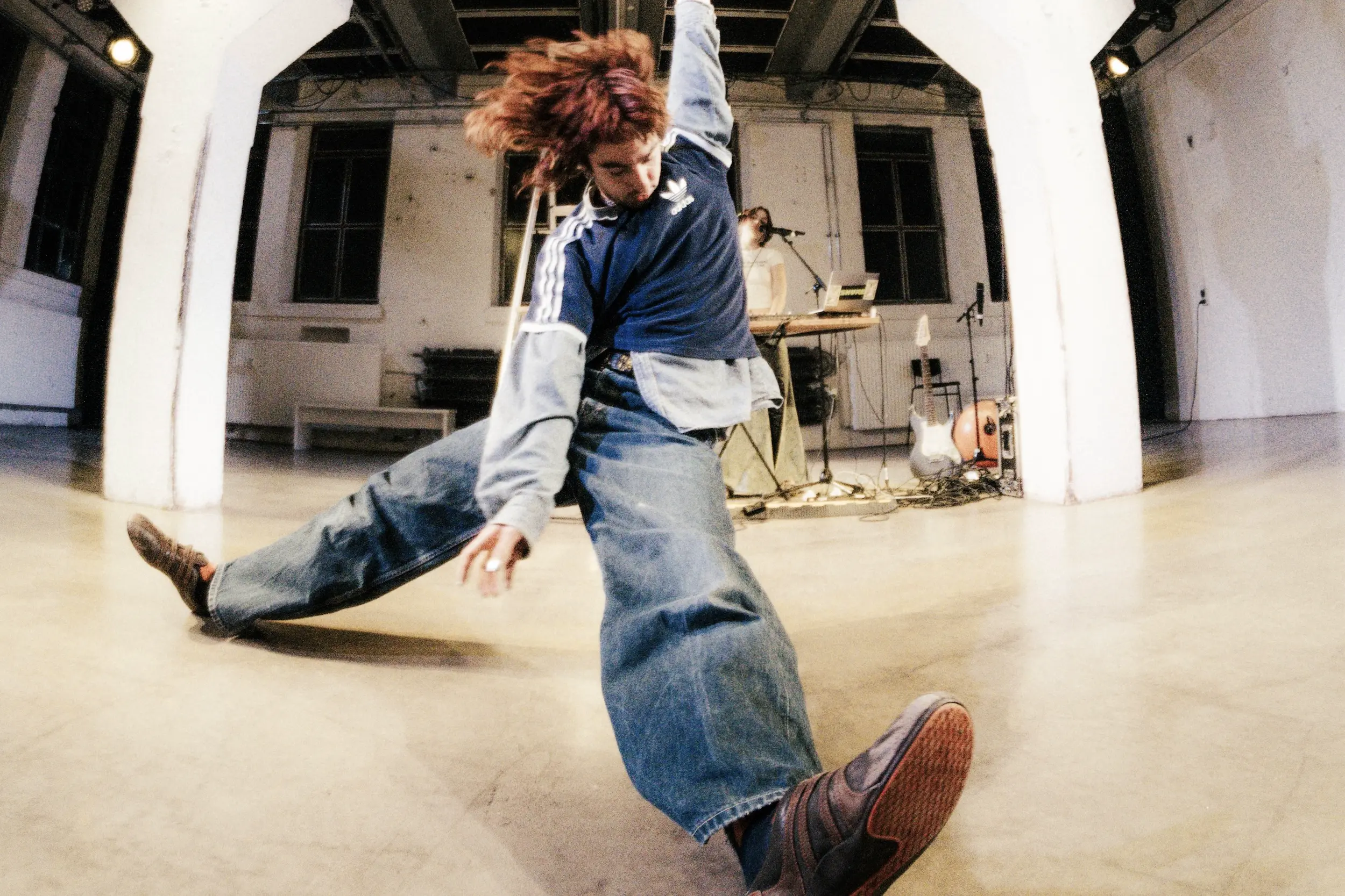 Tanzende Person in weitem Jeans-Outfit, die mit gespreizten Beinen und ausgestrecktem Arm über den Bühnenboden rutscht, hinter ihr stehen Instrumente, Technik und eine Performer:in am Mischpult.