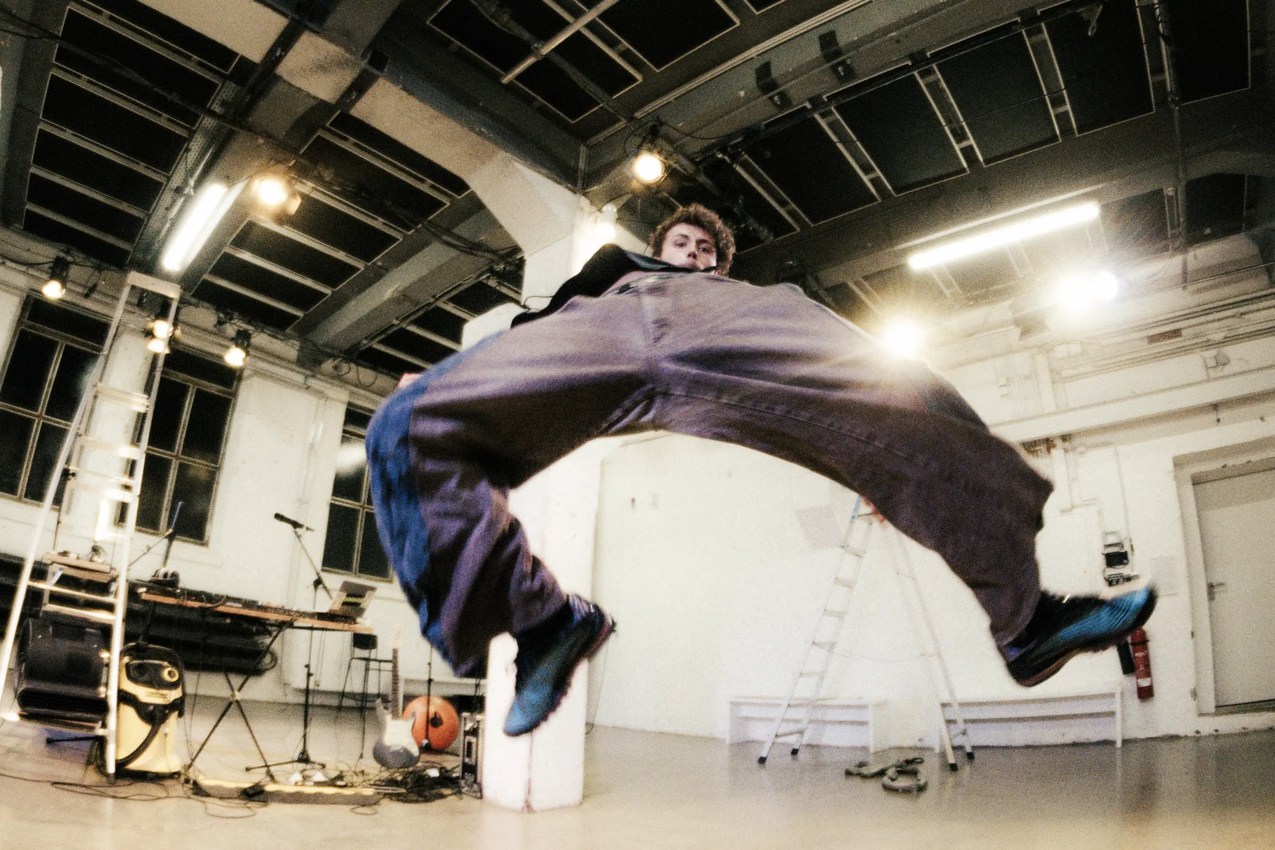 Weitwinkelaufnahme eines Sprungs: Eine Person springt mit weit gespreizten Beinen hoch in die Luft, sodass der Körper einen Bogen über der Kamera bildet, im Hintergrund der kahle Probebühnenraum mit Leitern und Scheinwerfern.