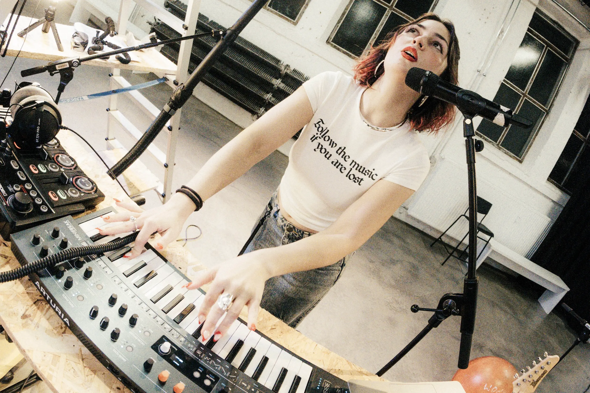 Nahaufnahme einer Musiker:in an einem Synthesizer, die gleichzeitig spielt und in ein Mikrofon singt; sie trägt ein T-Shirt mit Schriftzug, im Hintergrund ein leerer Probenraum mit Fenstern und Radiatoren.