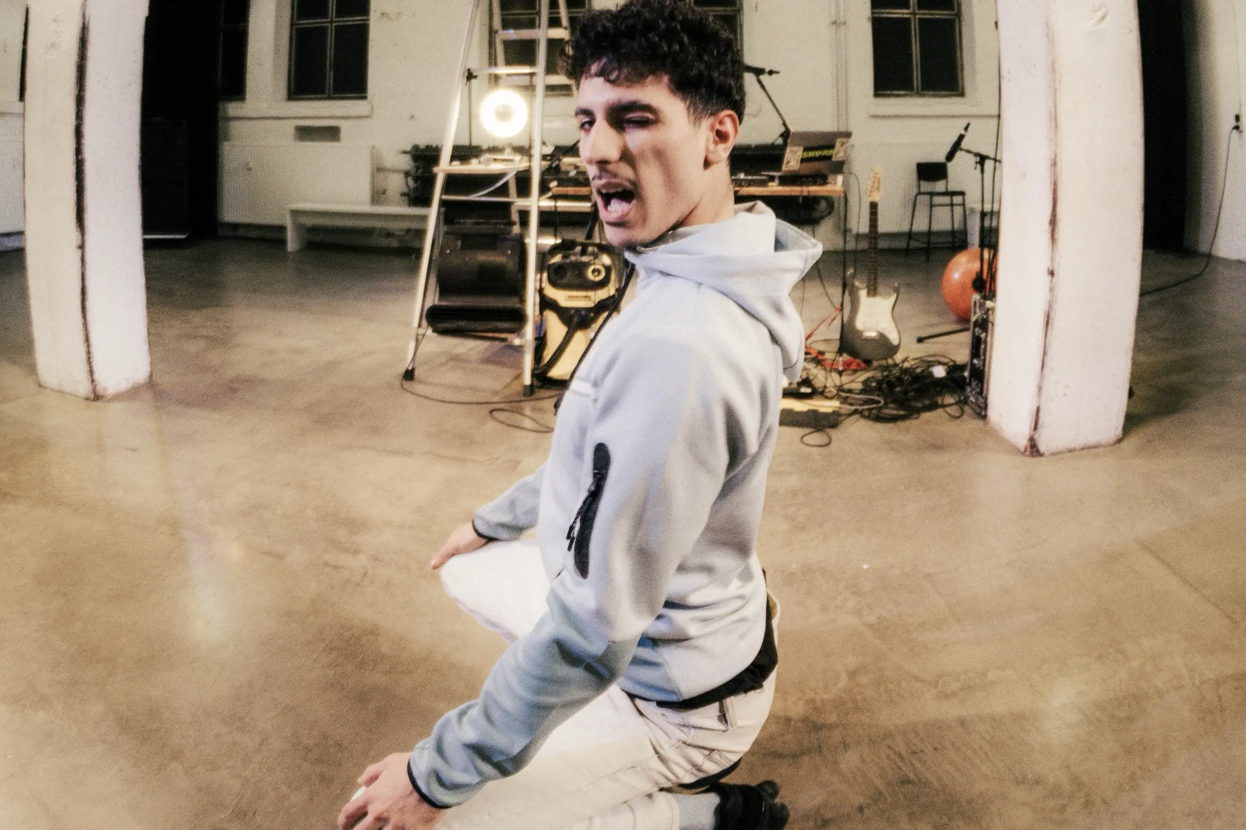 Ein Performer kniet mittig in einem leeren Bühnenraum, trägt einen hellen Hoodie, schaut zur Kamera und zieht das Gesicht in eine übertriebene Pose, im Hintergrund sind Technik, Instrumente und eine Leiter zu sehen.