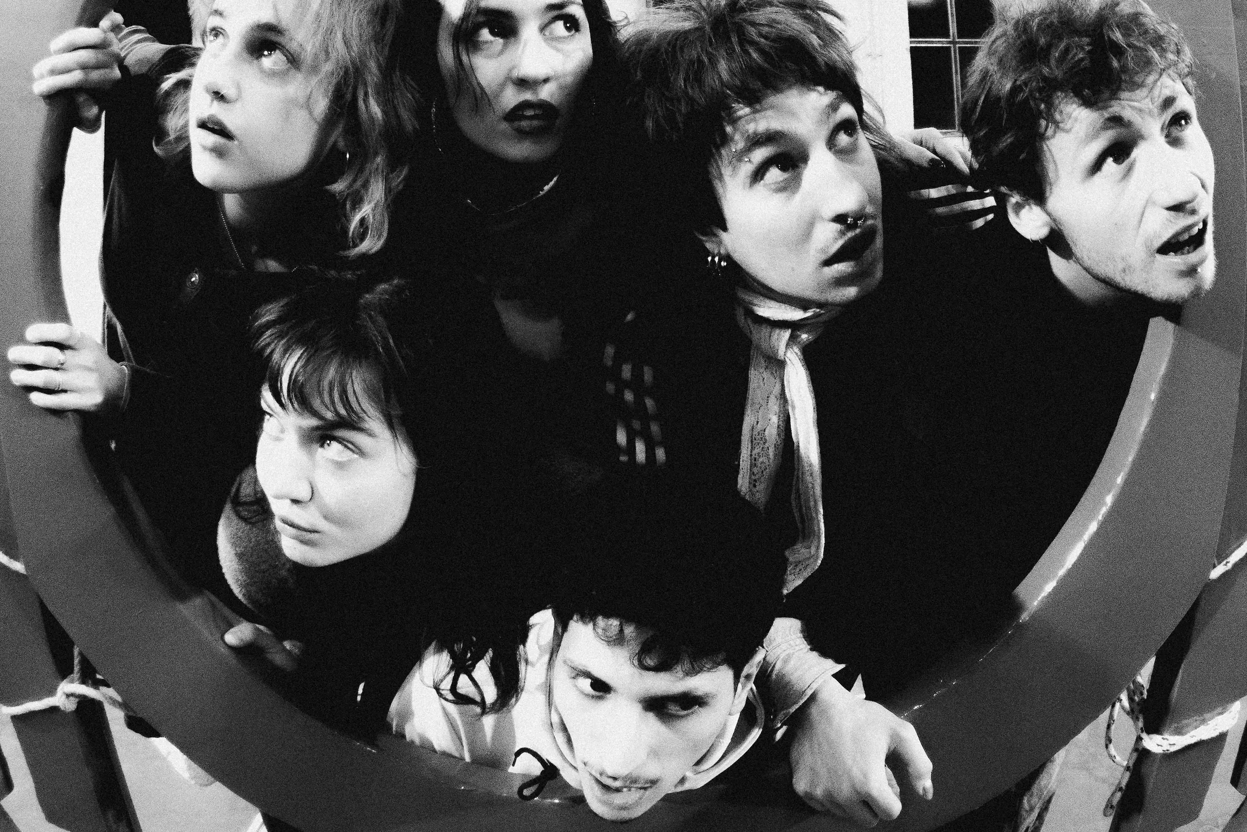 Schwarzweiß-Gruppenfoto von sechs jungen Menschen, die dicht gedrängt in einem runden Rahmen stehen und alle in unterschiedliche Richtungen nach oben blicken, als würden sie neugierig aus einer Luke schauen.