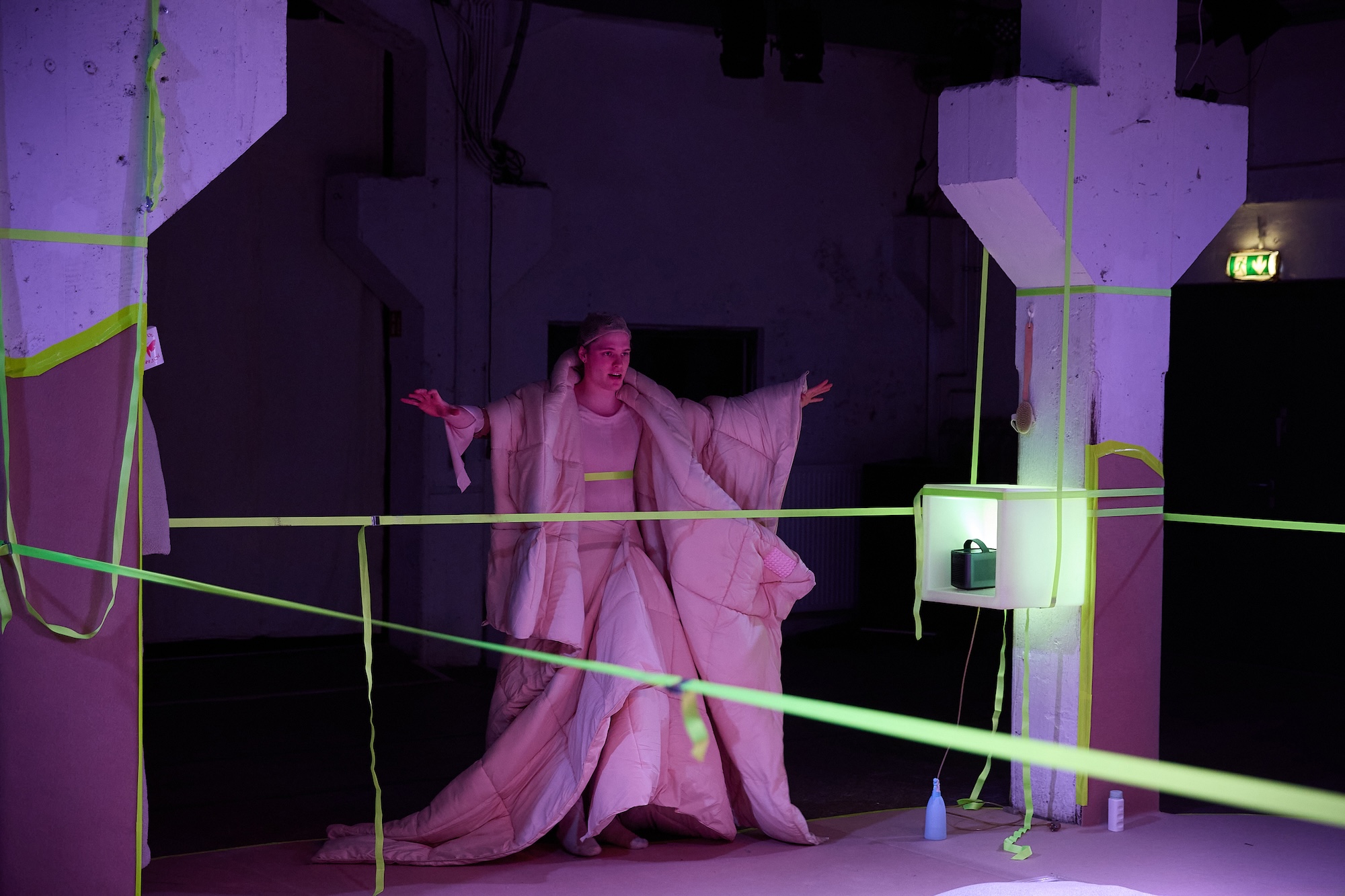 Eine Person in einem voluminösen, wattierten hellen Kostüm steht in einem ebenfalls von neongelben Seilen umspannten Bühnenraum, die Arme ausgebreitet, mit nachdenklichem Gesichtsausdruck. Die Szene ist in violettes und grünes Licht getaucht.