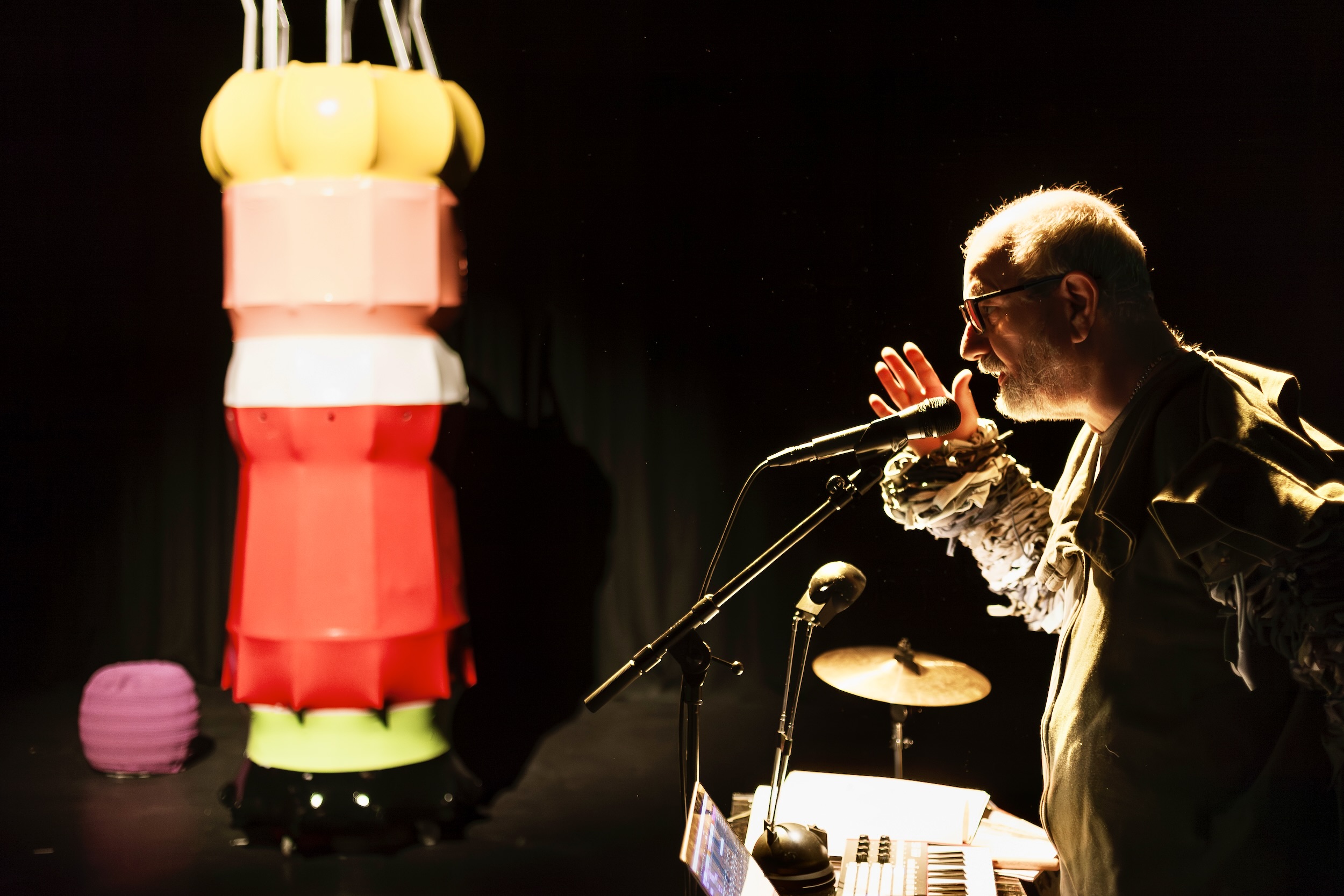 Person an einem Mikrofon auf dunkler Bühne neben einer überdimensionierten Strickliesel.