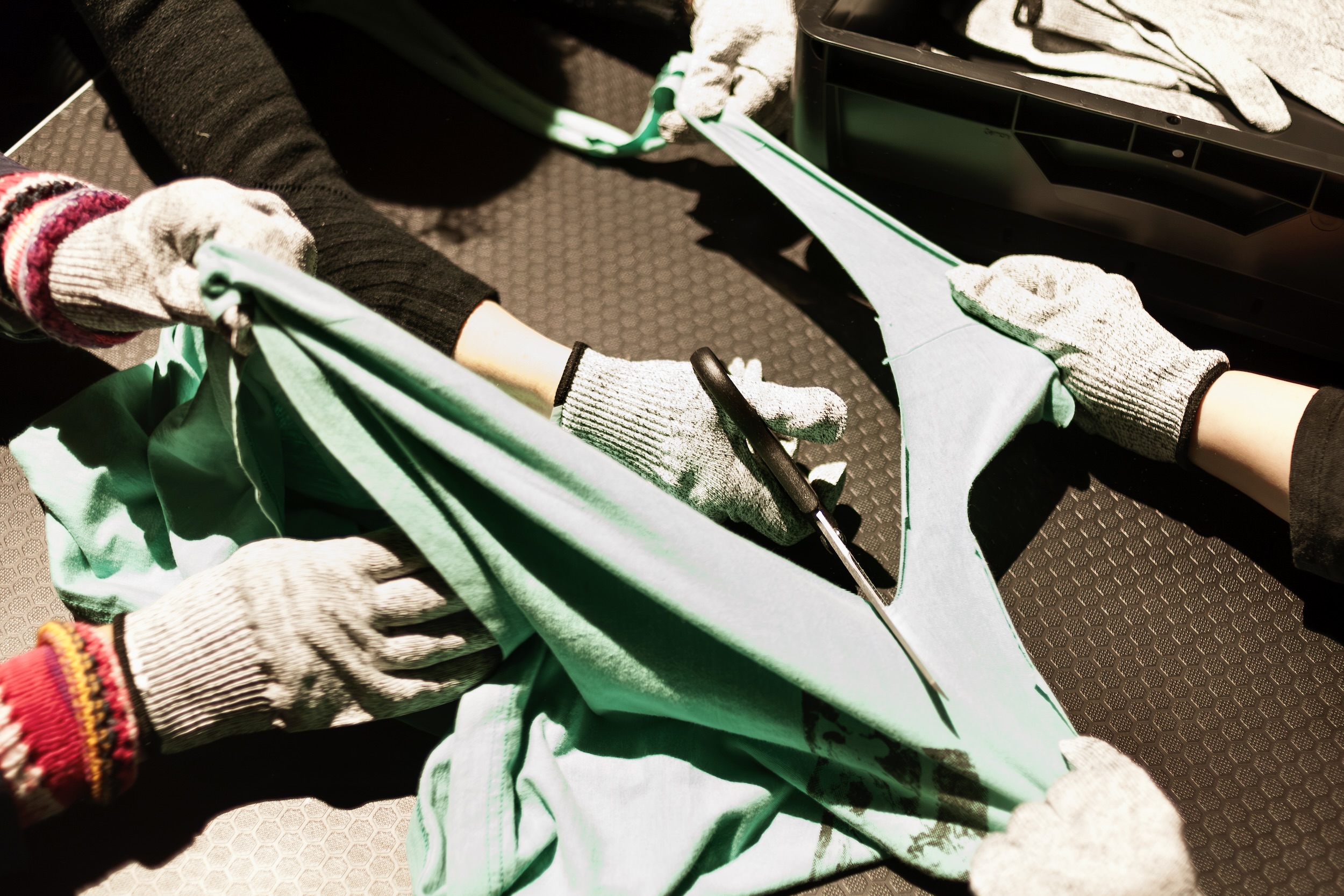 Mehrere behandschuhte Hände ziehen an Stoffen und zerschneiden ein Kleidungsstück