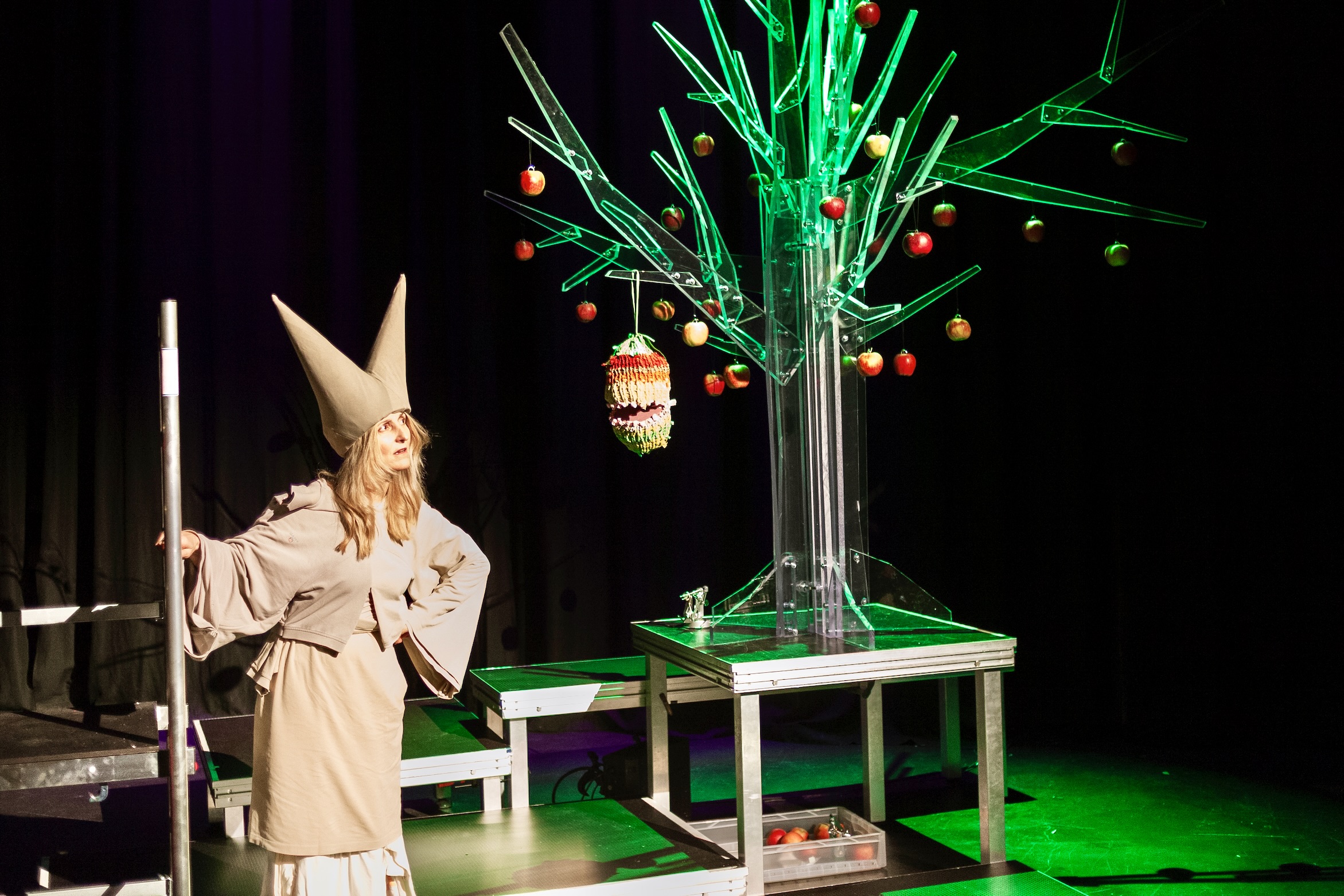 Person in hellem, spitzem Hut und weitem Gewand steht auf Bühne vor leuchtendem Kunstbaum mit hängenden Äpfeln.
