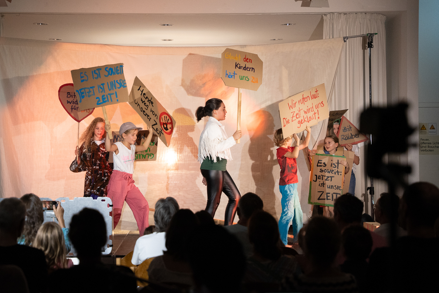 Eine Gruppe von Kindern demonstriert auf einer Theaterbühne mit selbst gebastelten Protestschildern, auf denen Slogans rund um Zeit und Kindheit geschrieben stehen. Vor der Bühne sitzt ein Publikum.
