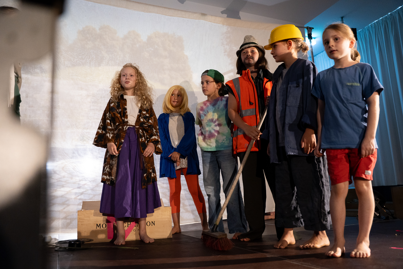 Sechs Kinder stehen in verschiedenen bunten Kostümen und ohne Schuhe auf einer Bühne, blicken nach vorne, im Hintergrund ist eine Leinwand mit Landschaftsprojektion zu sehen.