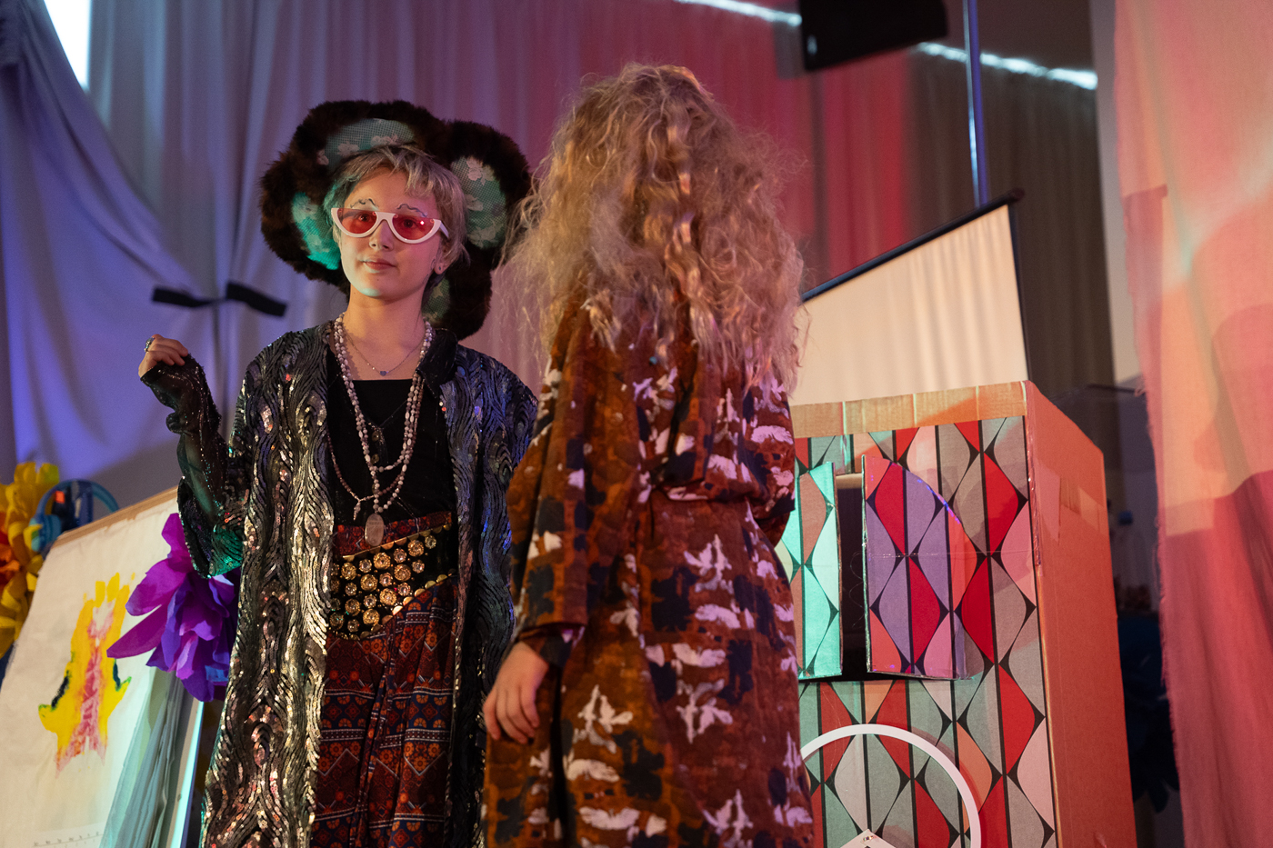 Zwei Kinder in fantasievollen Umhängen stehen sich auf einer bunt dekorierten Theaterbühne gegenüber, eine Kulisse mit Muster und Blumen ist im Hintergrund sichtbar.